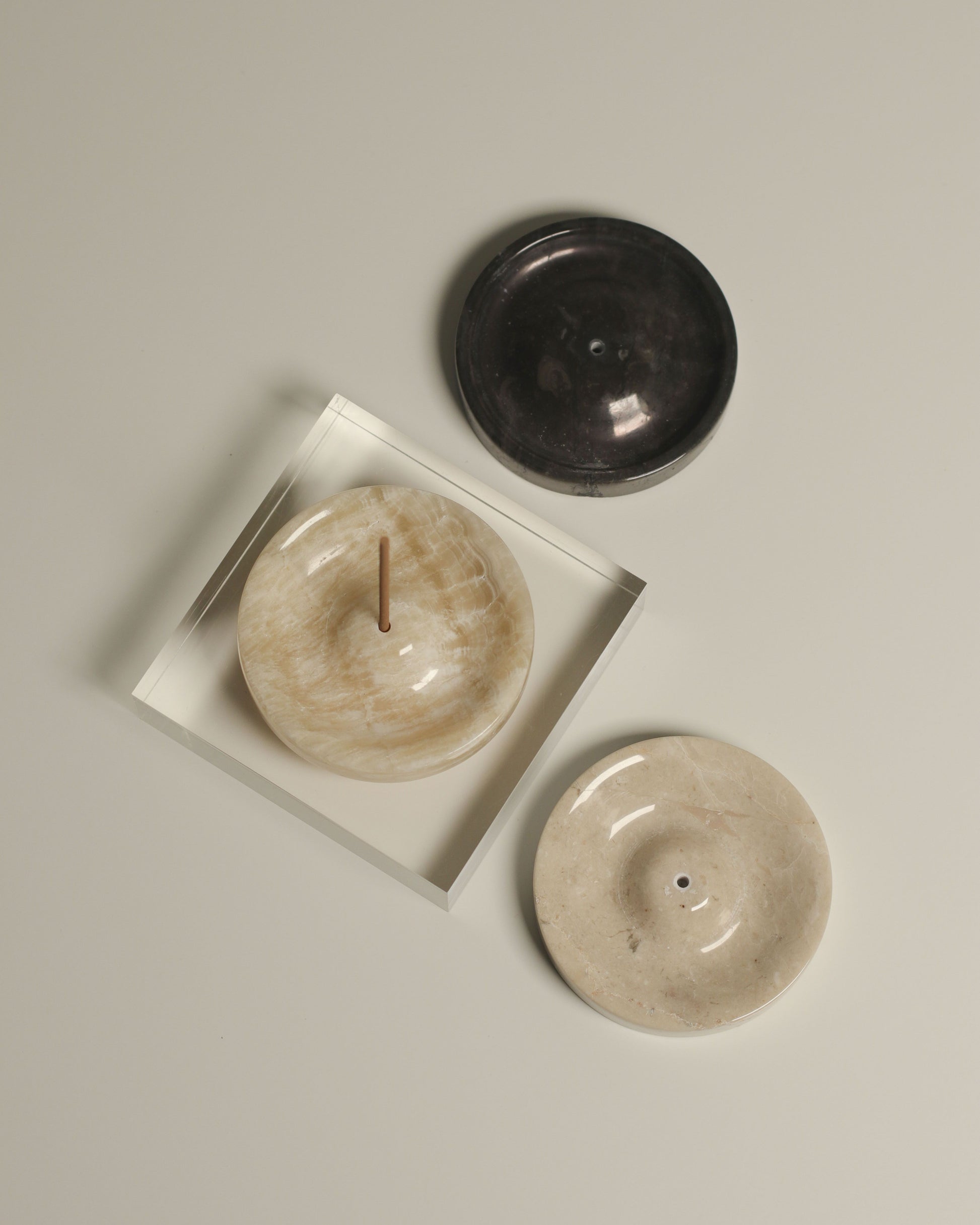 Three stillgoods Ebb Incense Holders in beige and dark gray are displayed on a light surface, with one placed inside a clear accessory tray holding an incense stick.