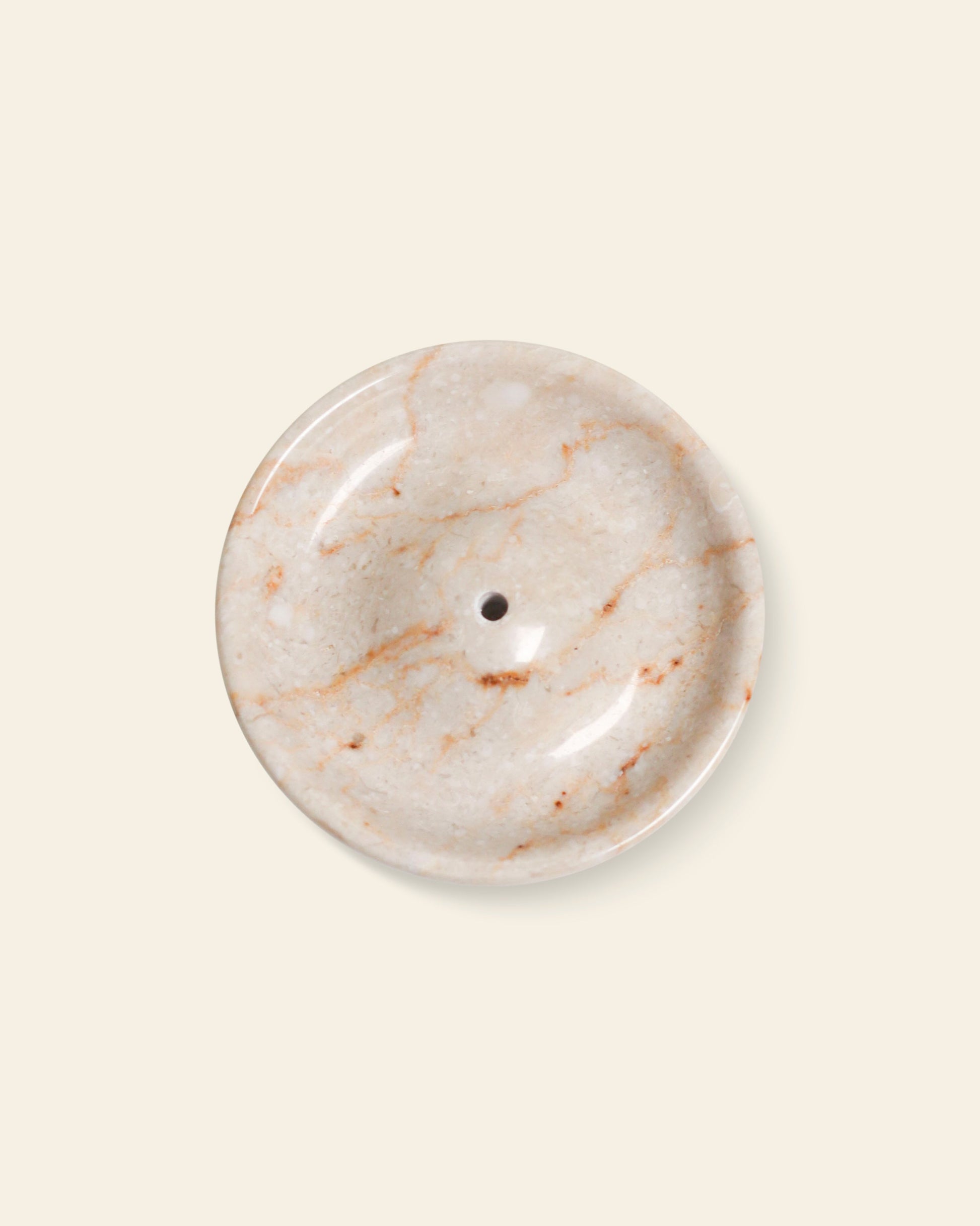The stillgoods Ebb Incense Holder by stillgoods is a round, beige marble tray with subtle orange-brown veining and a central drainage hole, shown on a plain light background.
