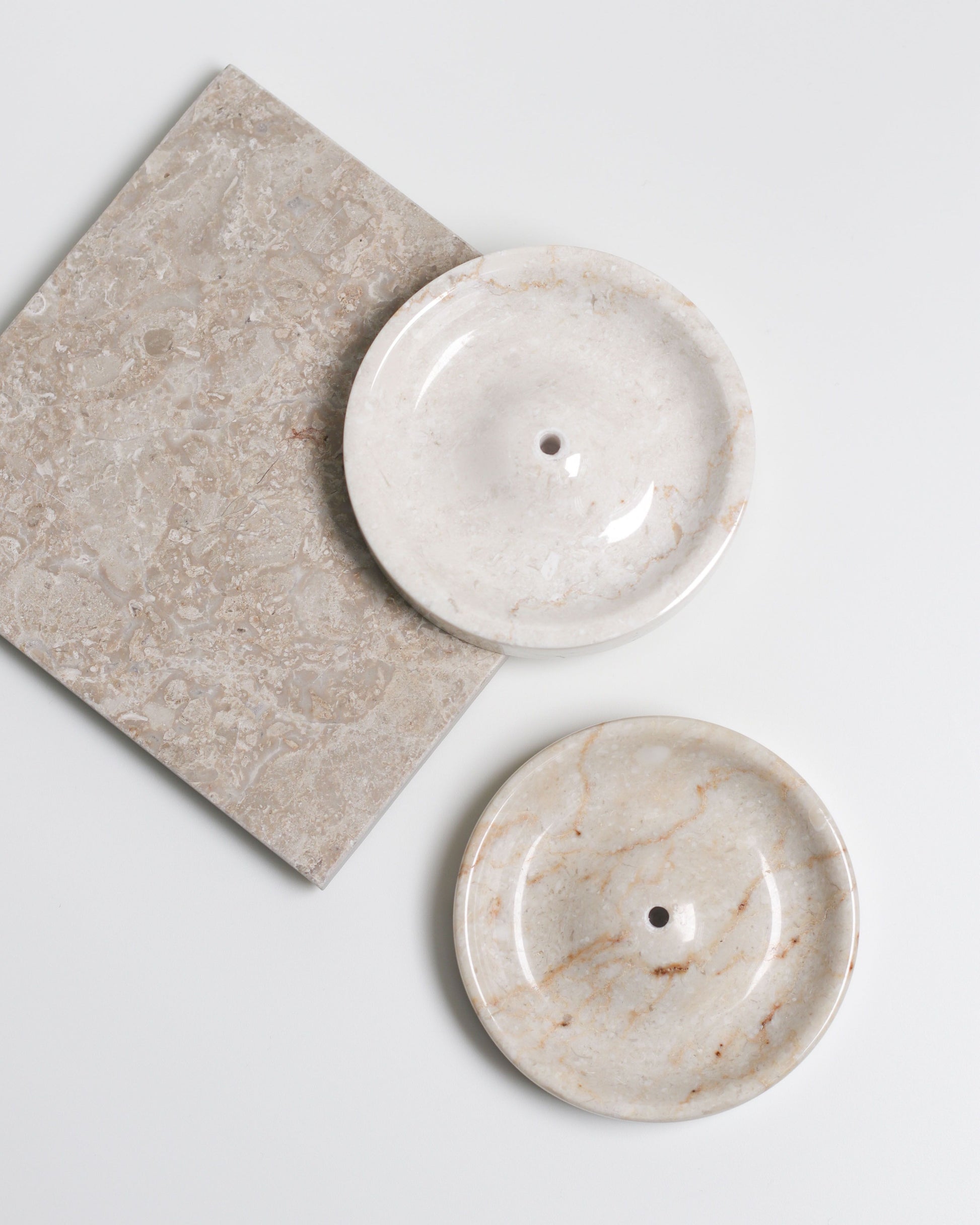 Two stillgoods Ebb Incense Holders—round, cream-colored marble with central drainage holes—are shown on and beside a square neutral stone tile, all against a white background. Versatile for use as an accessory tray or incense holder.