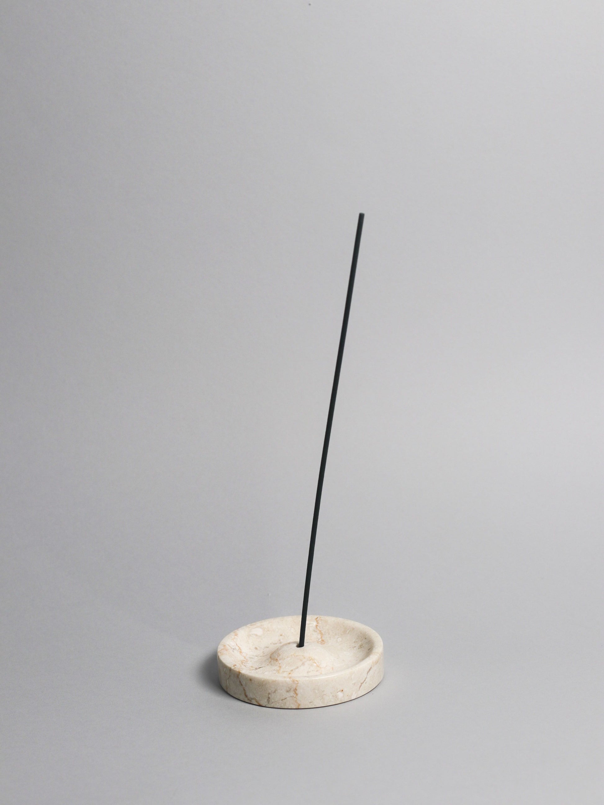 A black incense stick stands upright in the stillgoods Ebb Incense Holder, a round marble design by stillgoods, against a plain light gray background.