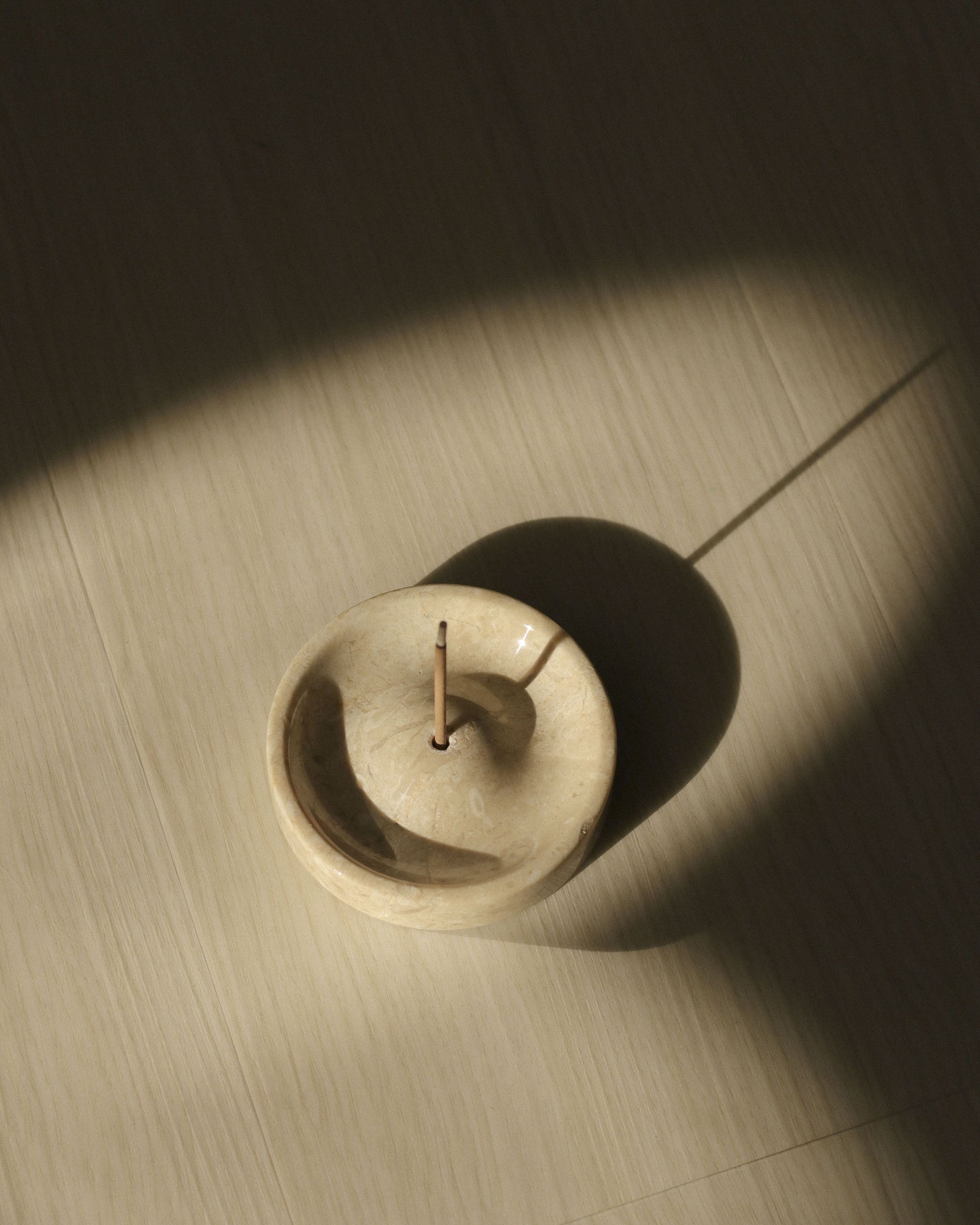 The stillgoods Ebb Incense Holder by stillgoods, shown here with a burning incense stick, casts a gentle shadow on a sunlit wooden floor. Its smooth beige surface is perfect as an accessory tray or ikebana holder.