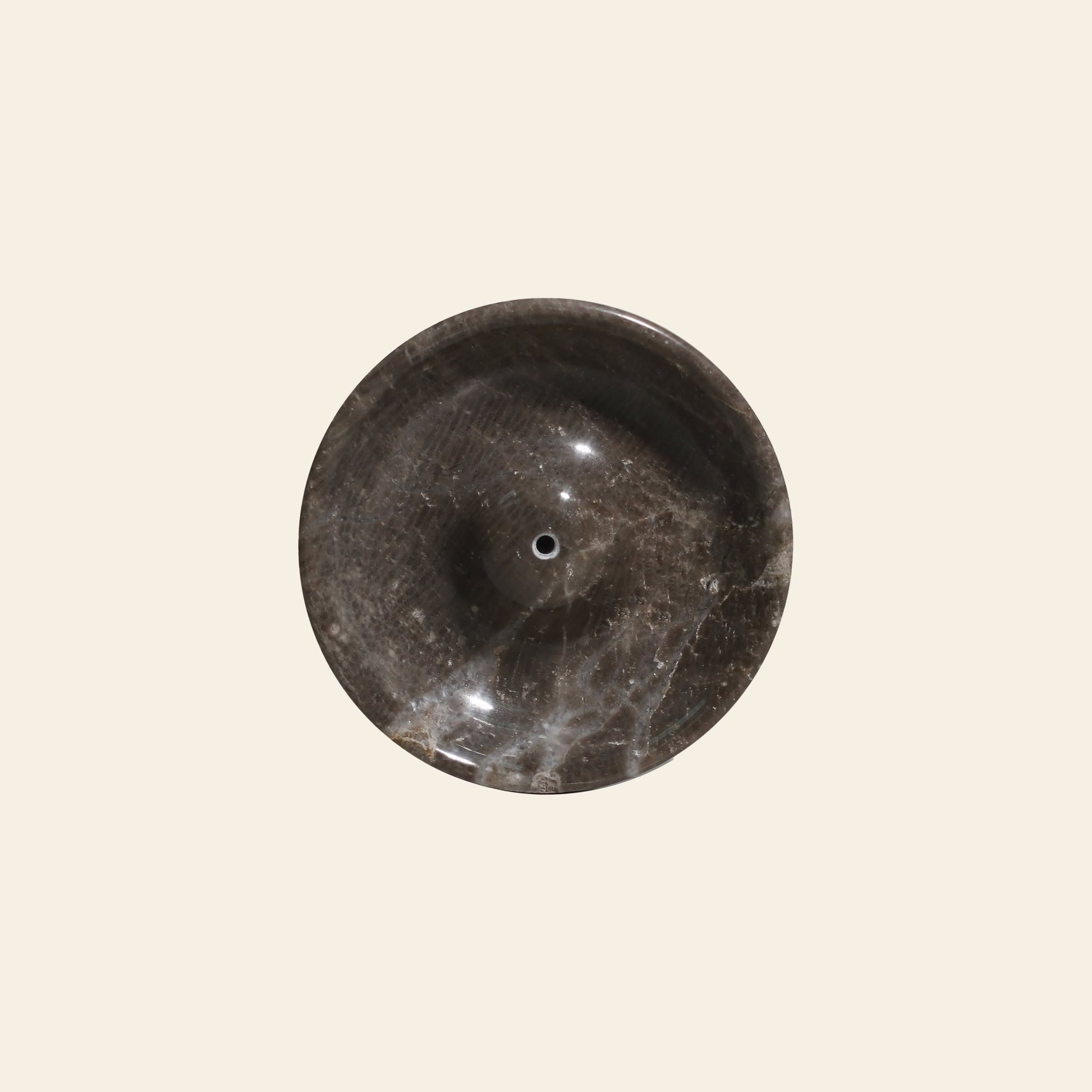 The stillgoods Ebb Incense Holder features a round, dark marble design with white veining, glossy finish, and petite drain hole. Centered on a plain light beige background, it brings modern elegance to your space.