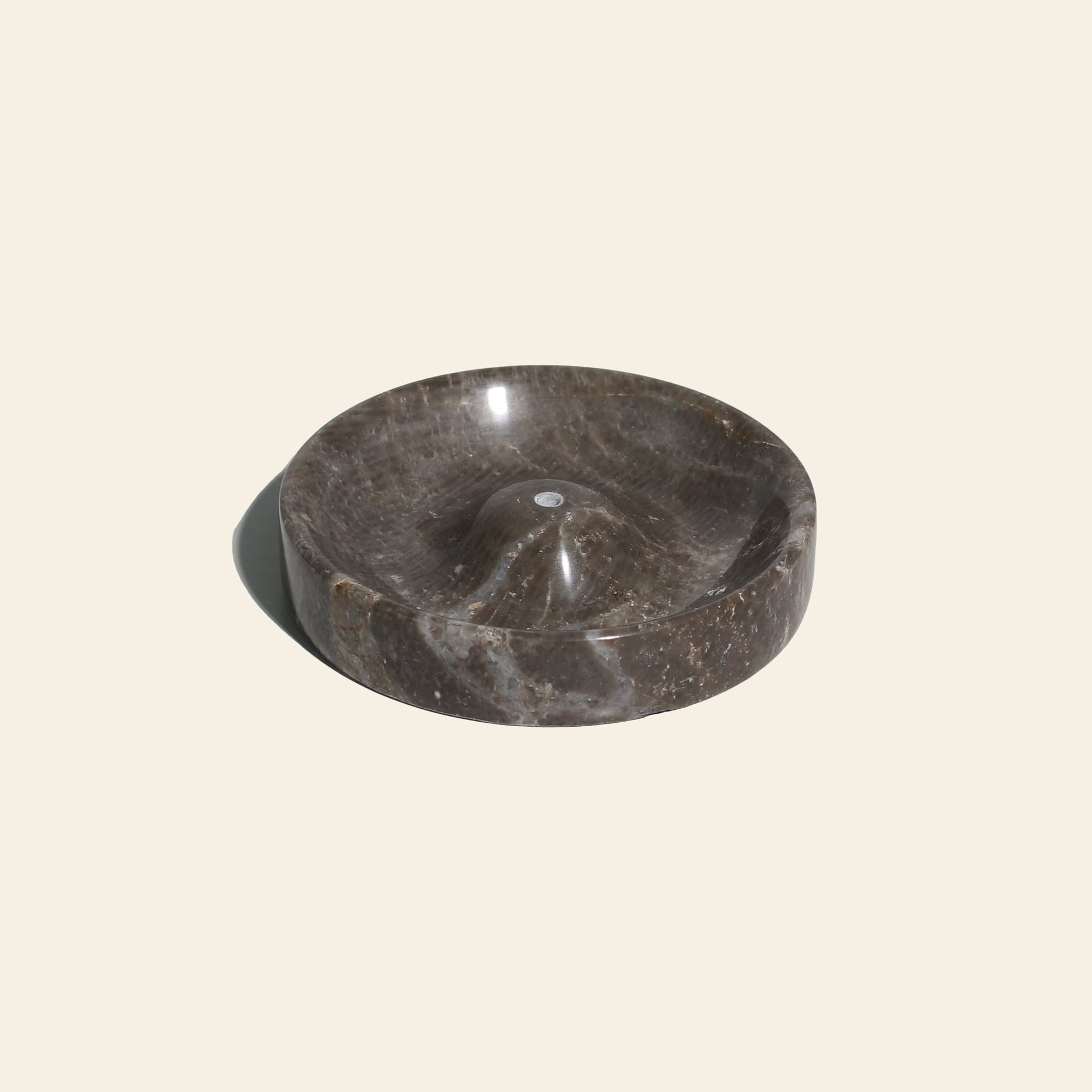 The stillgoods Ebb Incense Holder is a round, dark brown marble tray featuring a central raised area and a small groove, shown on a plain light beige background.