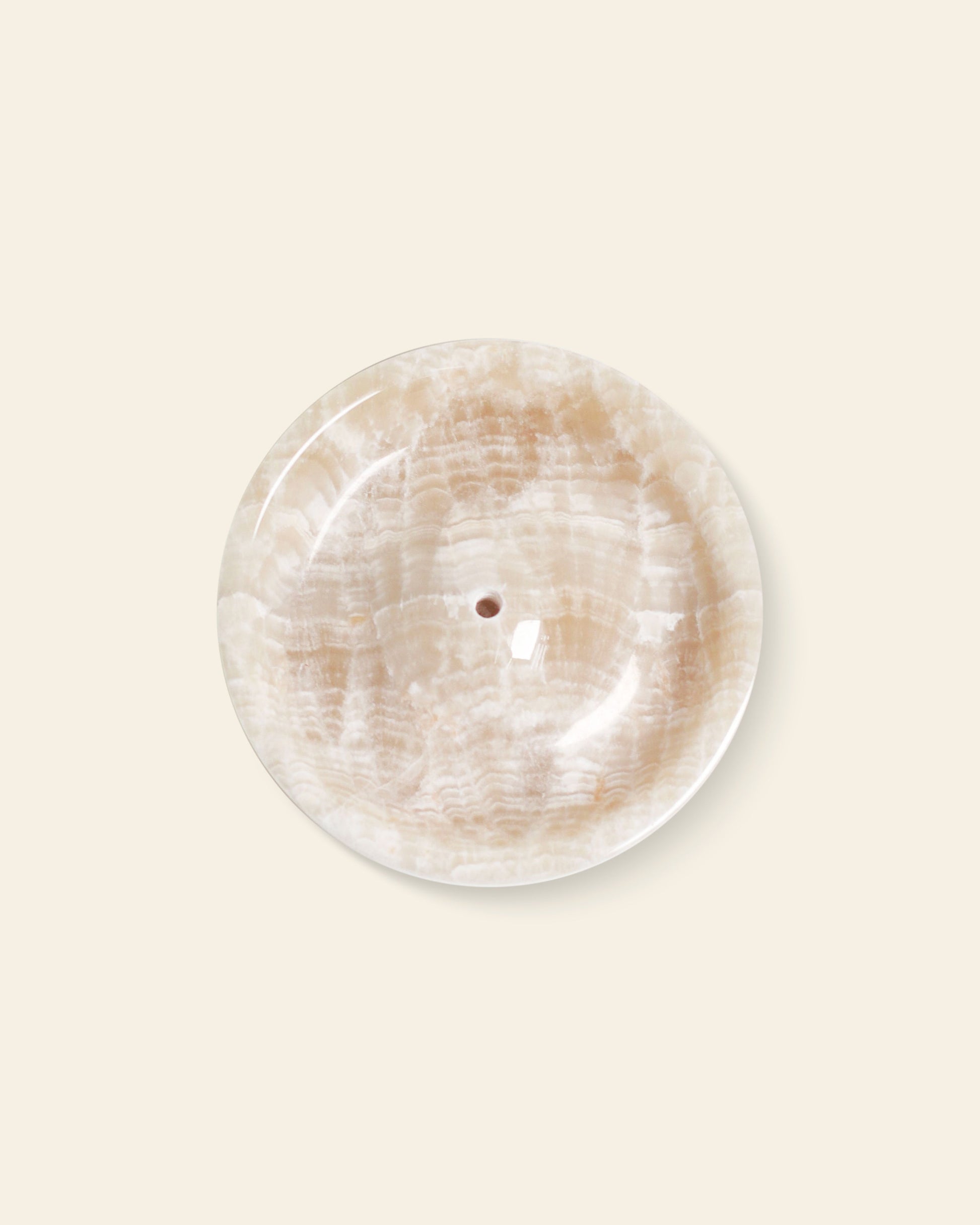 The stillgoods Ebb Incense Holder is a round, polished beige marble tray with white veining and a central drainage hole, ideal as an incense holder or elegant decor piece. Shown against a light cream background.