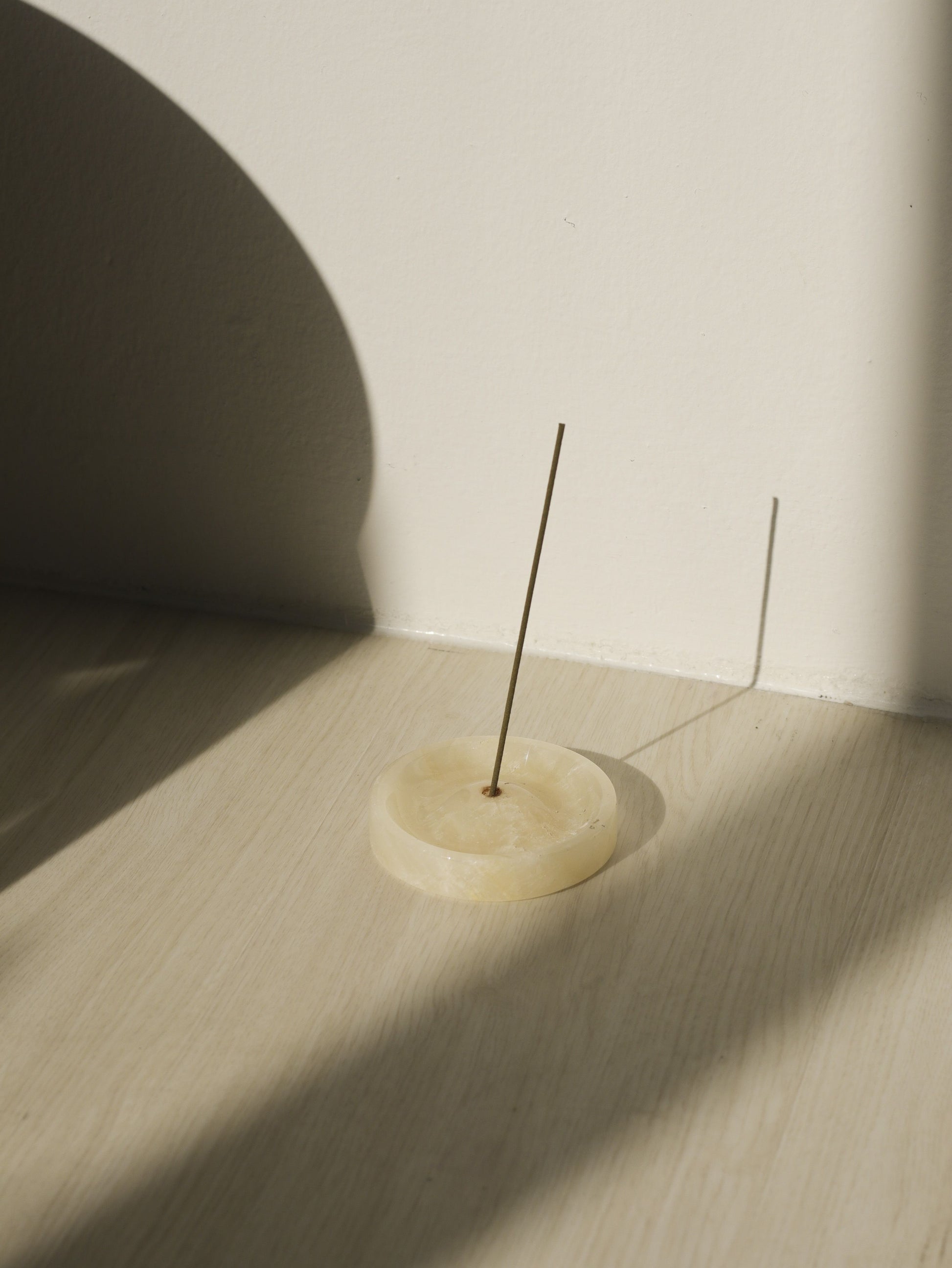 A single incense stick stands upright in the stillgoods Ebb Incense Holder—a round, cream-colored piece—on a light wooden floor, with sunlight casting soft shadows on the floor and wall.