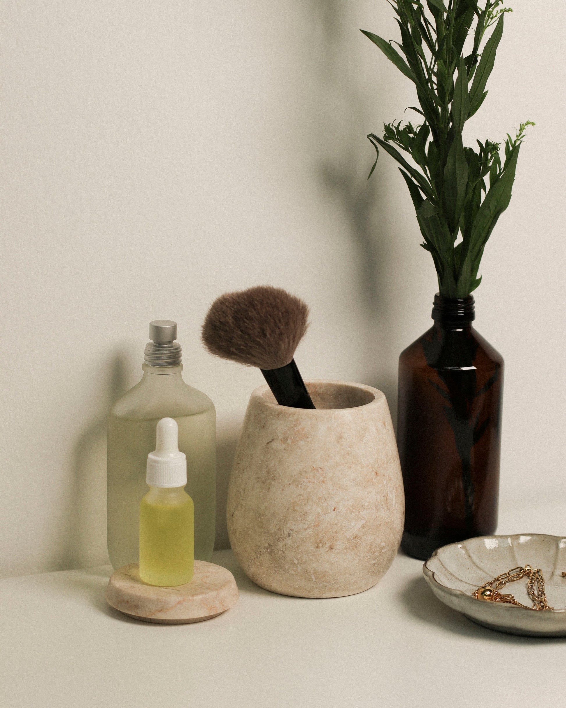 A glass dropper bottle with yellow liquid, a frosted spray bottle, the stillgoods Ecru Jar by stillgoods as an organizer, a makeup brush in a stone holder, and a dark vase with green leaves rest on a white surface against a light wall.