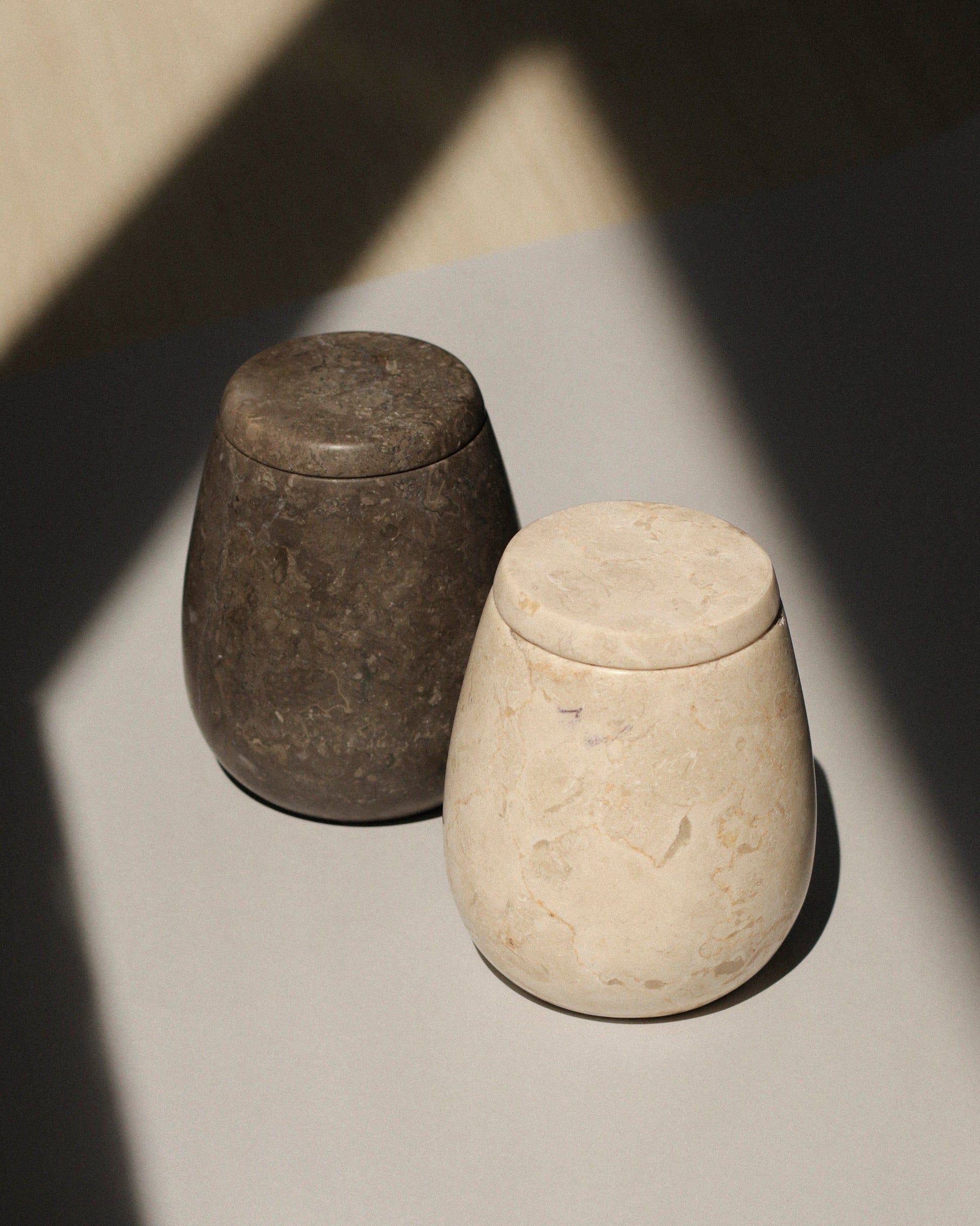 The stillgoods Ecru Jar and a dark gray stone jar sit side by side on a light surface, casting soft shadows. Ideal for organizing or storing condiments, these elegant jars from stillgoods enhance any setting with their refined style.