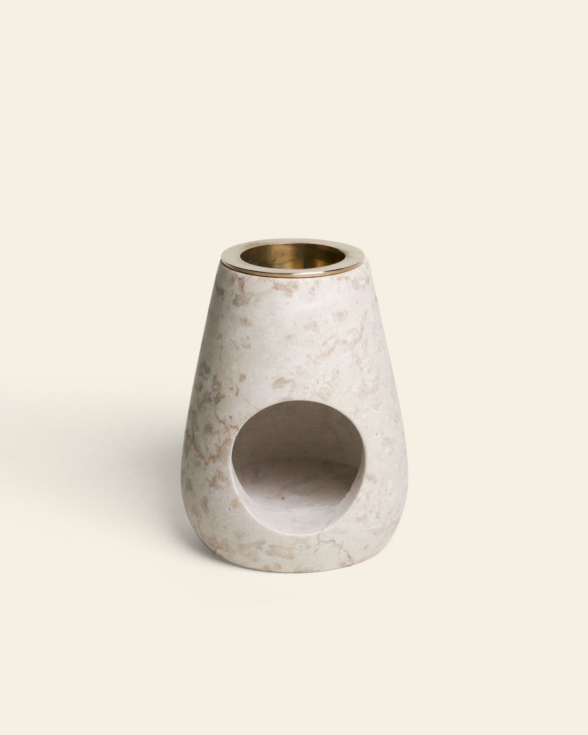 The stillgoods Eli Oil Burner is a conical, light marble diffuser with a brass insert and front circular opening, designed to fill your space with calming scents. Shown against a plain cream background.