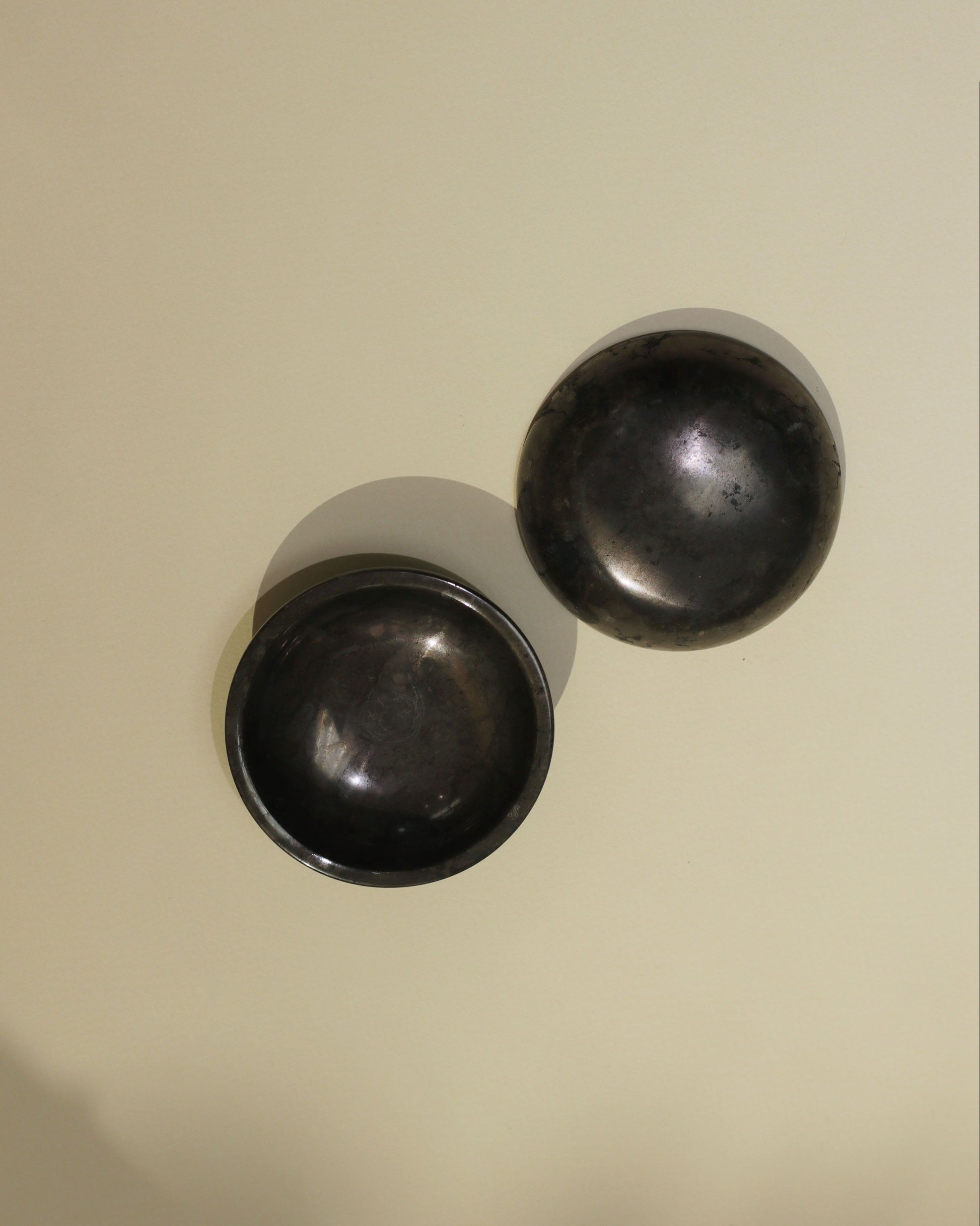 Two round stillgoods Luna Dishes with a dark, metallic black marble finish sit on a light beige surface. One lies flat while the other is angled on its side, both casting soft shadows.