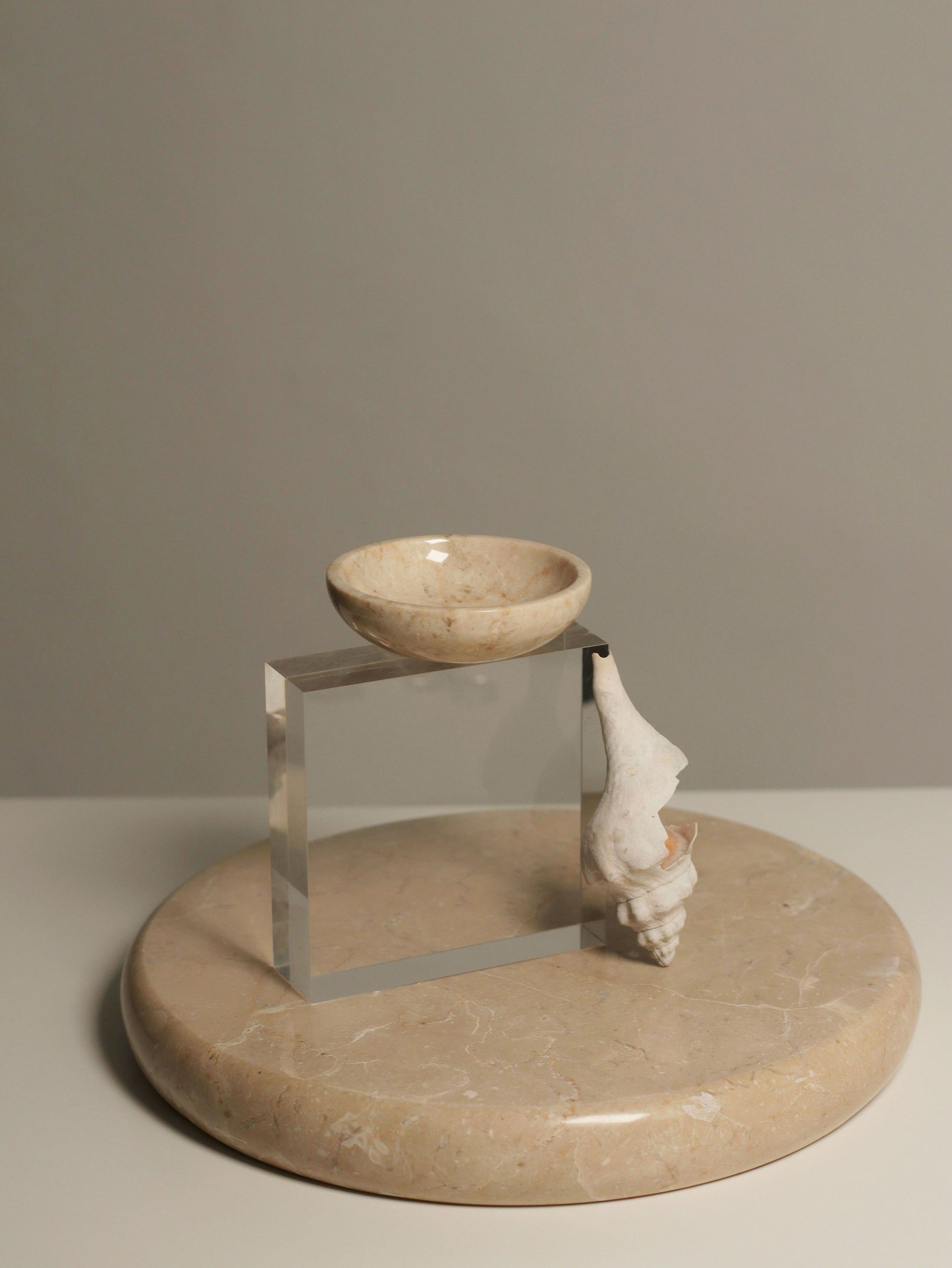 The stillgoods Luna Dish, a small marble catch-all from stillgoods, is displayed with a clear acrylic cube and a white seashell on a round beige marble platform against a plain gray background.