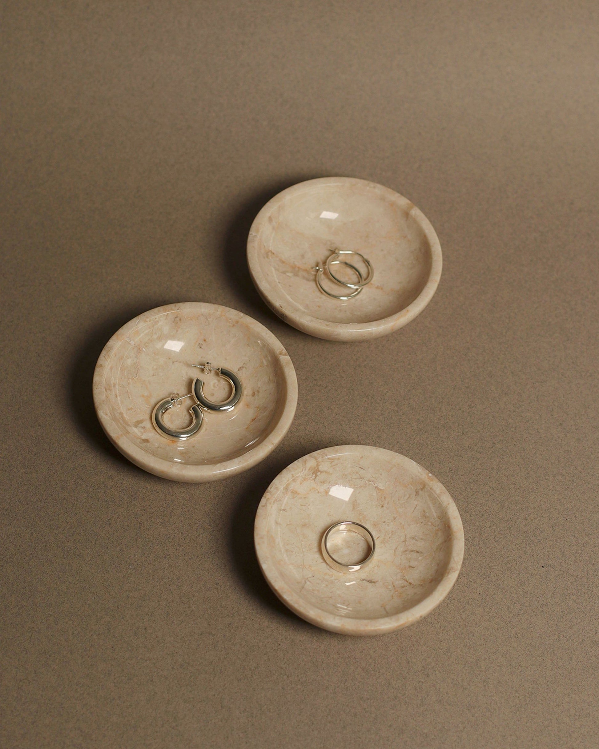 Three stillgoods Luna Dishes, small and round in beige marble, each display one or two silver rings on a smooth taupe surface.