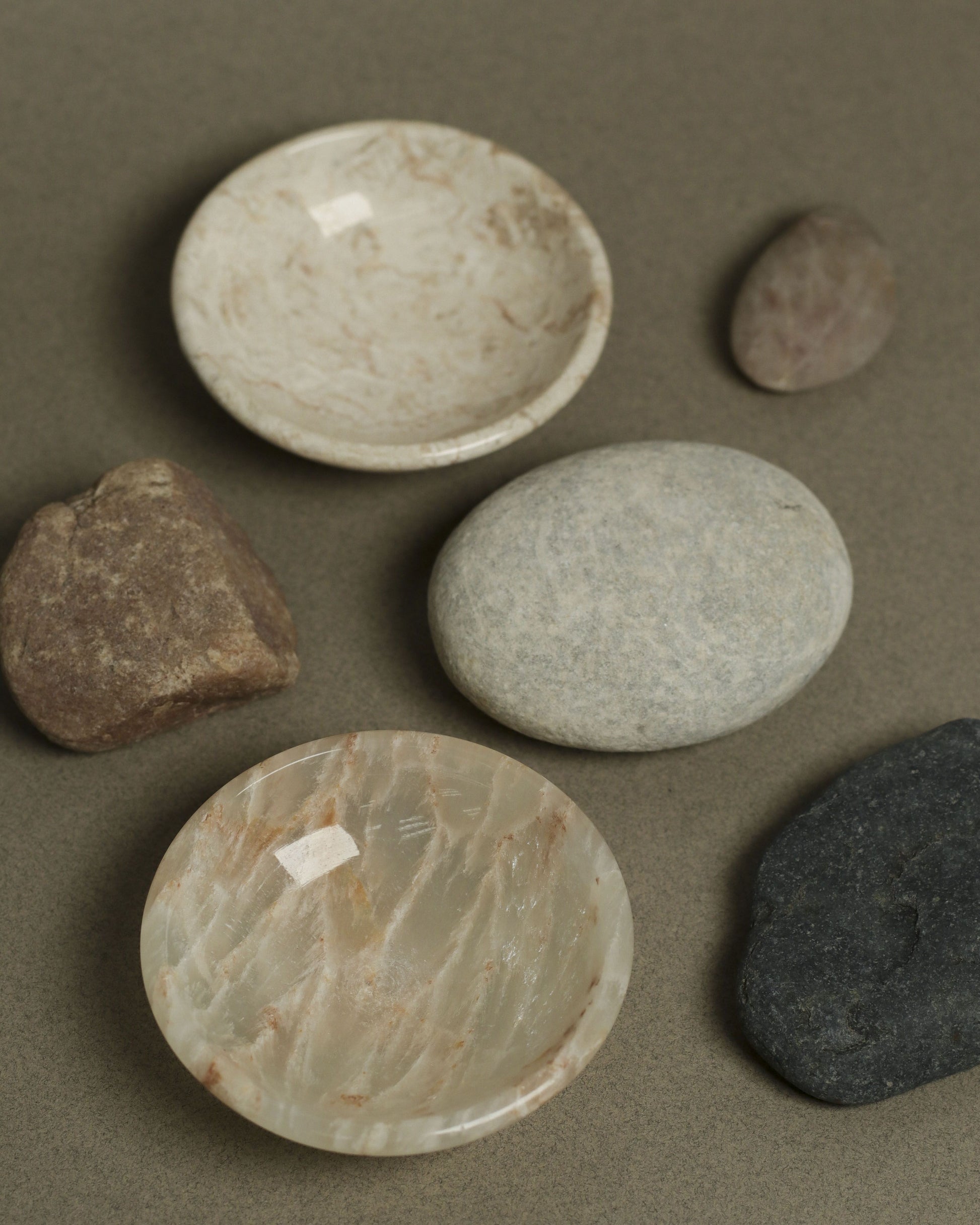 The stillgoods Luna Dish set by stillgoods includes two round marble bowls with natural beige patterns, ideal as trinket or catch-all dishes, displayed with four smooth, assorted stones on a neutral brown surface.