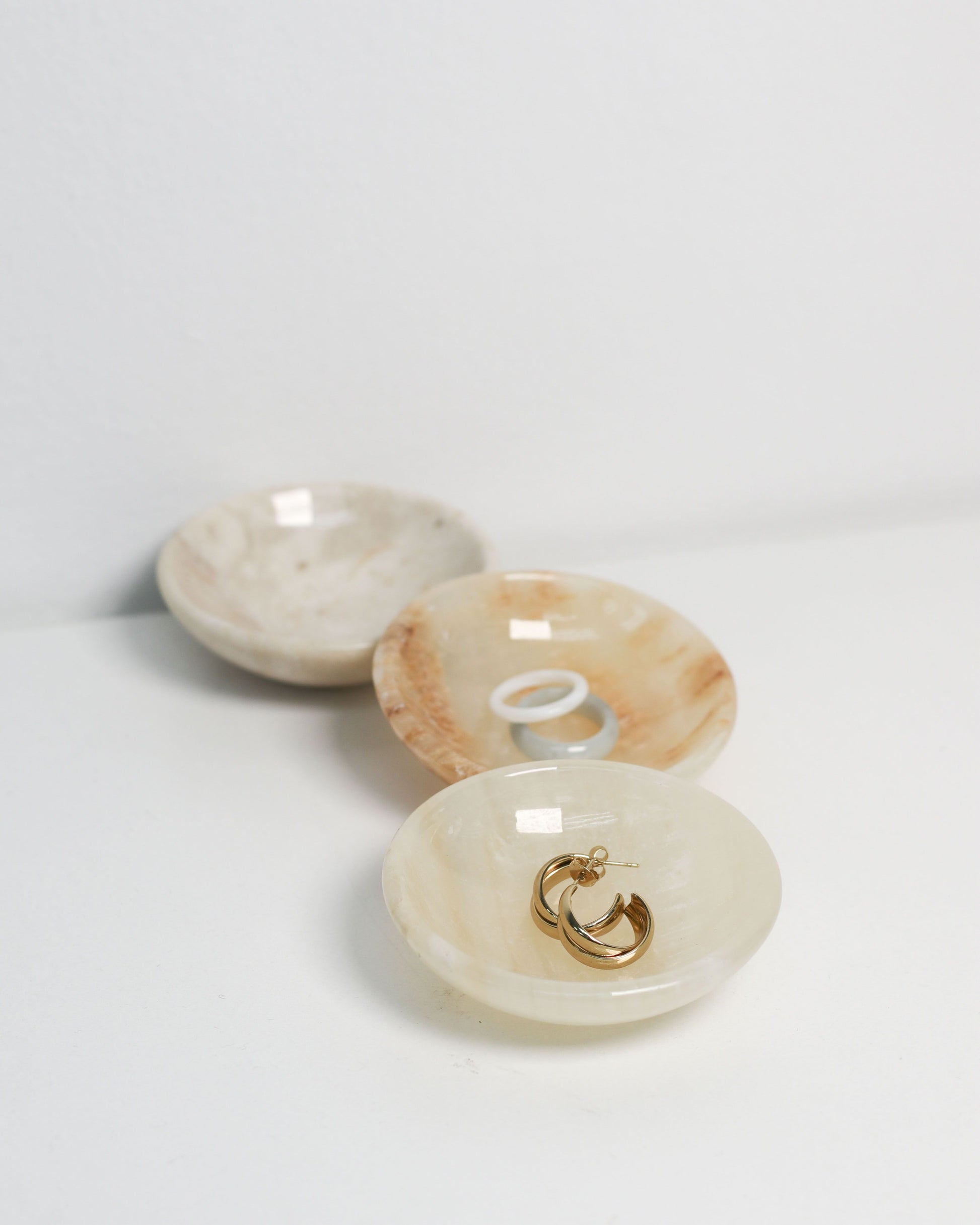 Three stillgoods Luna Dishes by stillgoods are arranged on a white surface: the front holds gold hoop earrings, the middle contains two rings, and the back dish is empty.