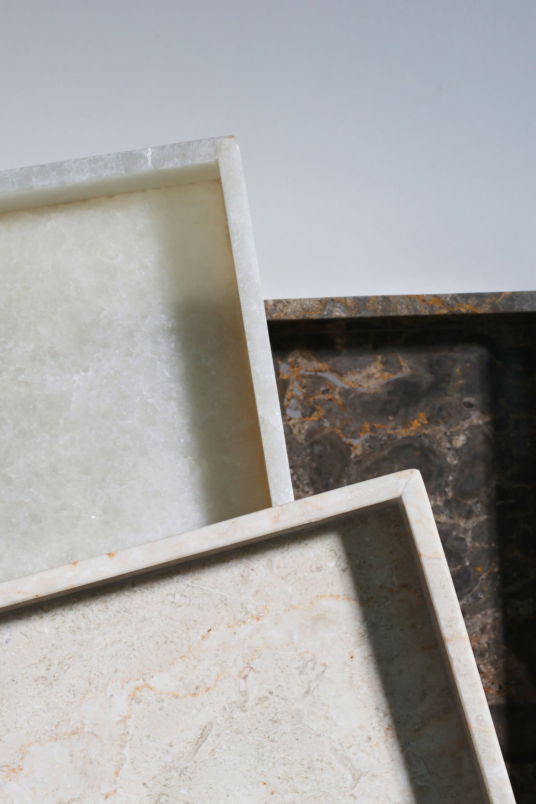 Three stillgoods stone trays—including the stillgoods Mini Valor Tray—in white, beige, and dark brown are stacked on a neutral background. Each rectangular tray features raised edges, low height, and a textured surface for organized essentials.