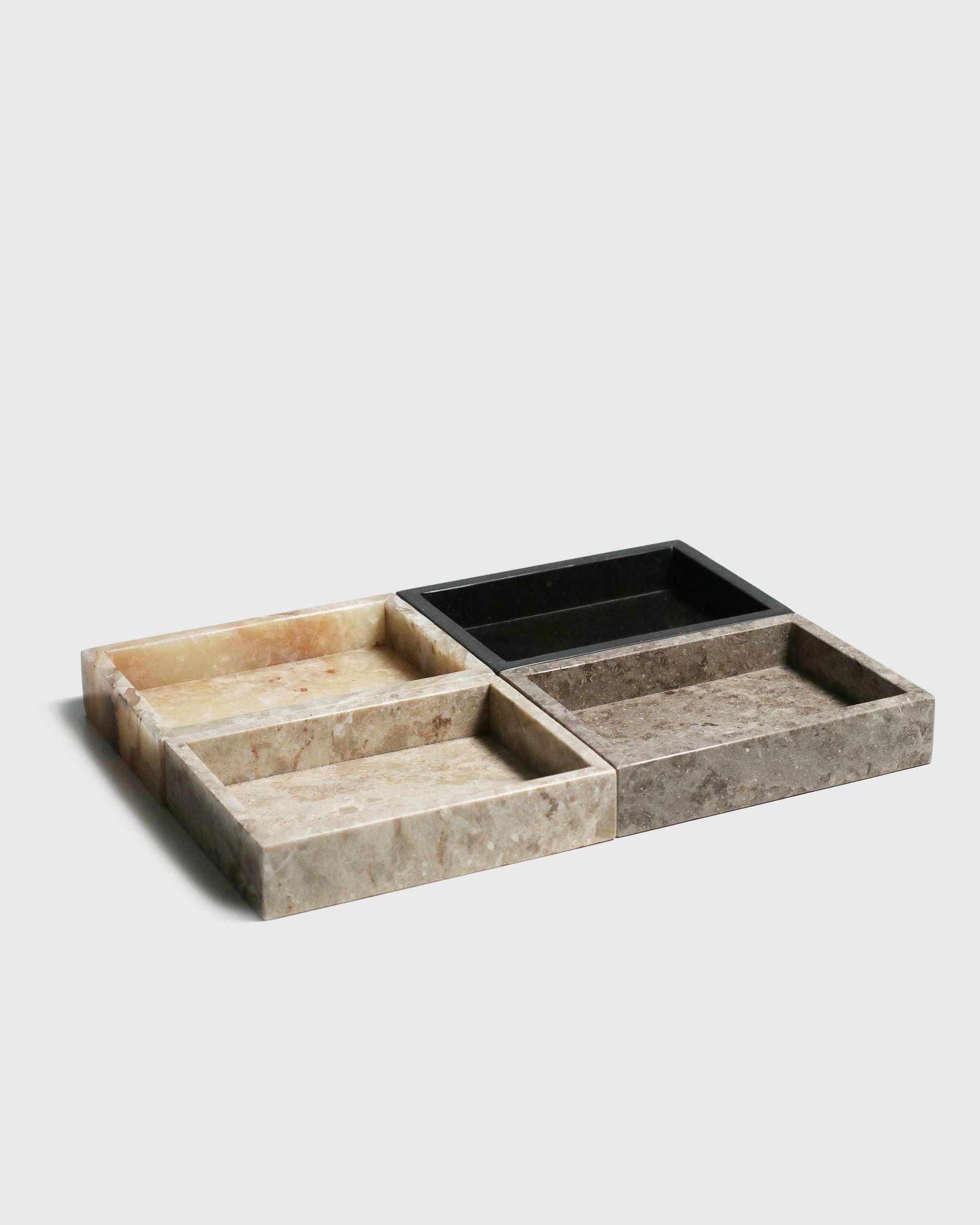 Four stillgoods Mini Valor Trays in beige, brown, and black are arranged on a light grey background. Each stillgoods tray has a low profile and polished finish—ideal for keeping essentials organized and stylishly displayed.