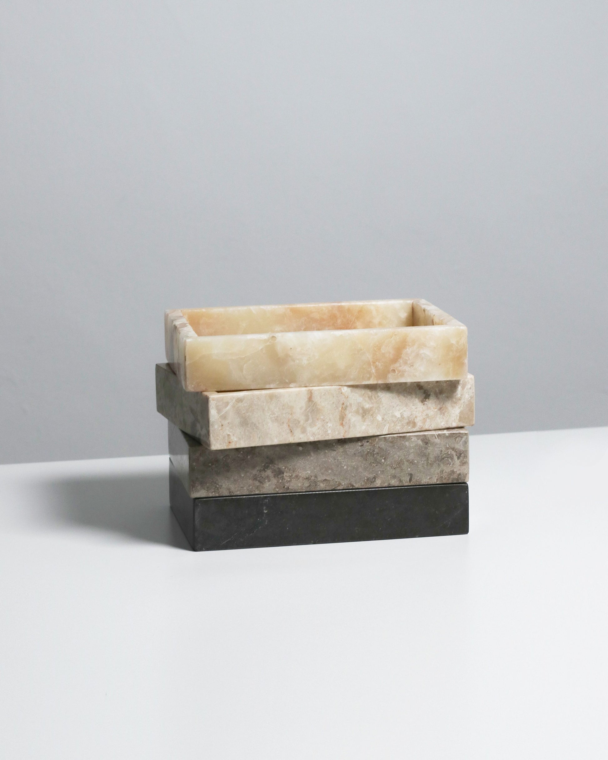 The stillgoods Mini Valor Trays—three rectangular stone trays in cream, beige, and dark gray—are stacked slightly off-center on a white surface, offering an elegant way to organize essentials against a light gray backdrop.