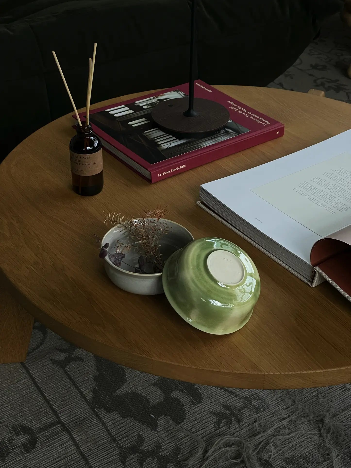 A wooden coffee table displays an open book, the ves Aki Bowl Set of 2 by ves inspired by East Asian tableware, a small dish with dried flowers, a reed diffuser, and a stack of books including a red hardcover in soft light.