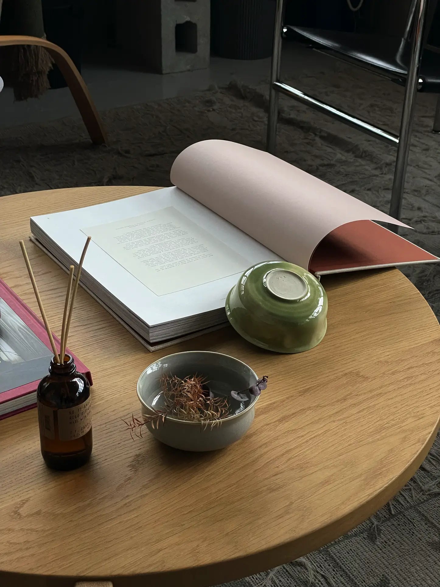 A handcrafted wooden table displays an open book, a green ves Aki Bowl (Set of 2) by ves—East Asian tableware—a small dish with dried plants, and a reed diffuser. A chair and textured carpet appear in the background.