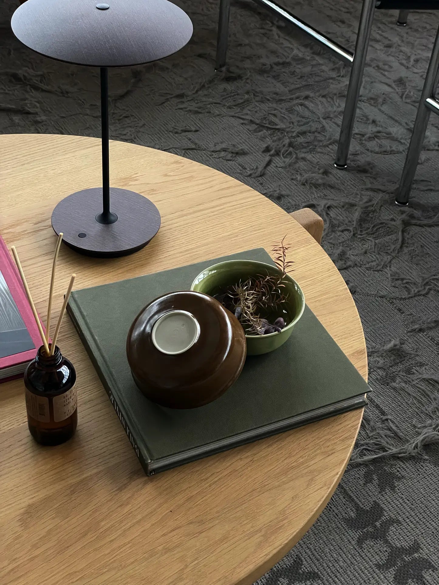 A round wooden table displays a green book, ves Aki Bowl (set of 2) by ves—one upside down—handcrafted East Asian tableware, a small green bowl with a dried plant, reed diffuser, and modern lamp atop a patterned carpet near chairs.