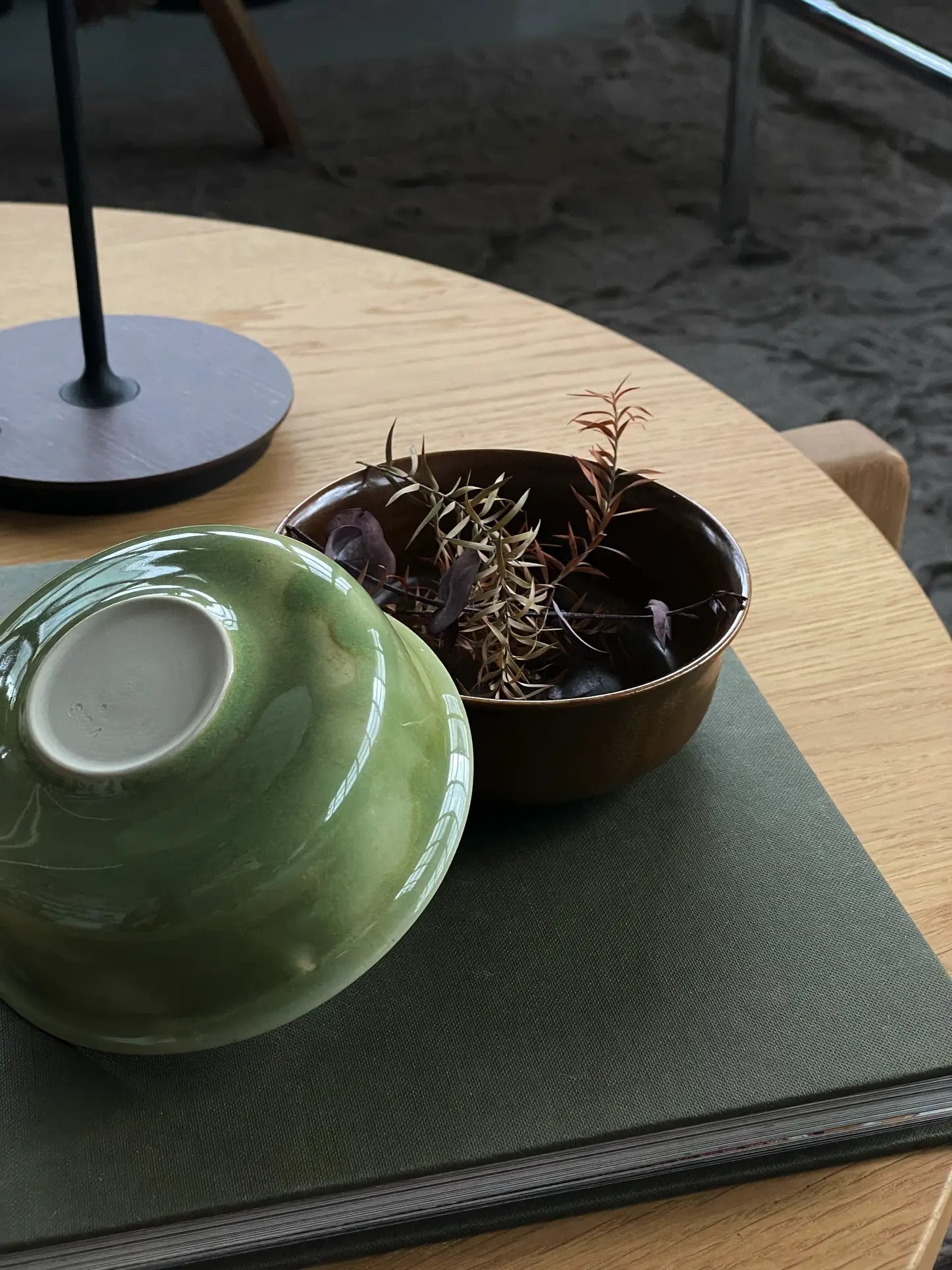 A ves Aki Bowl from the ves Aki Bowl, Set of 2 sits on a hardcover book atop a wooden table, beside an open brown bowl with dried spiky plants and purple leaves. In the background, a lamp base and sandy floor are visible.