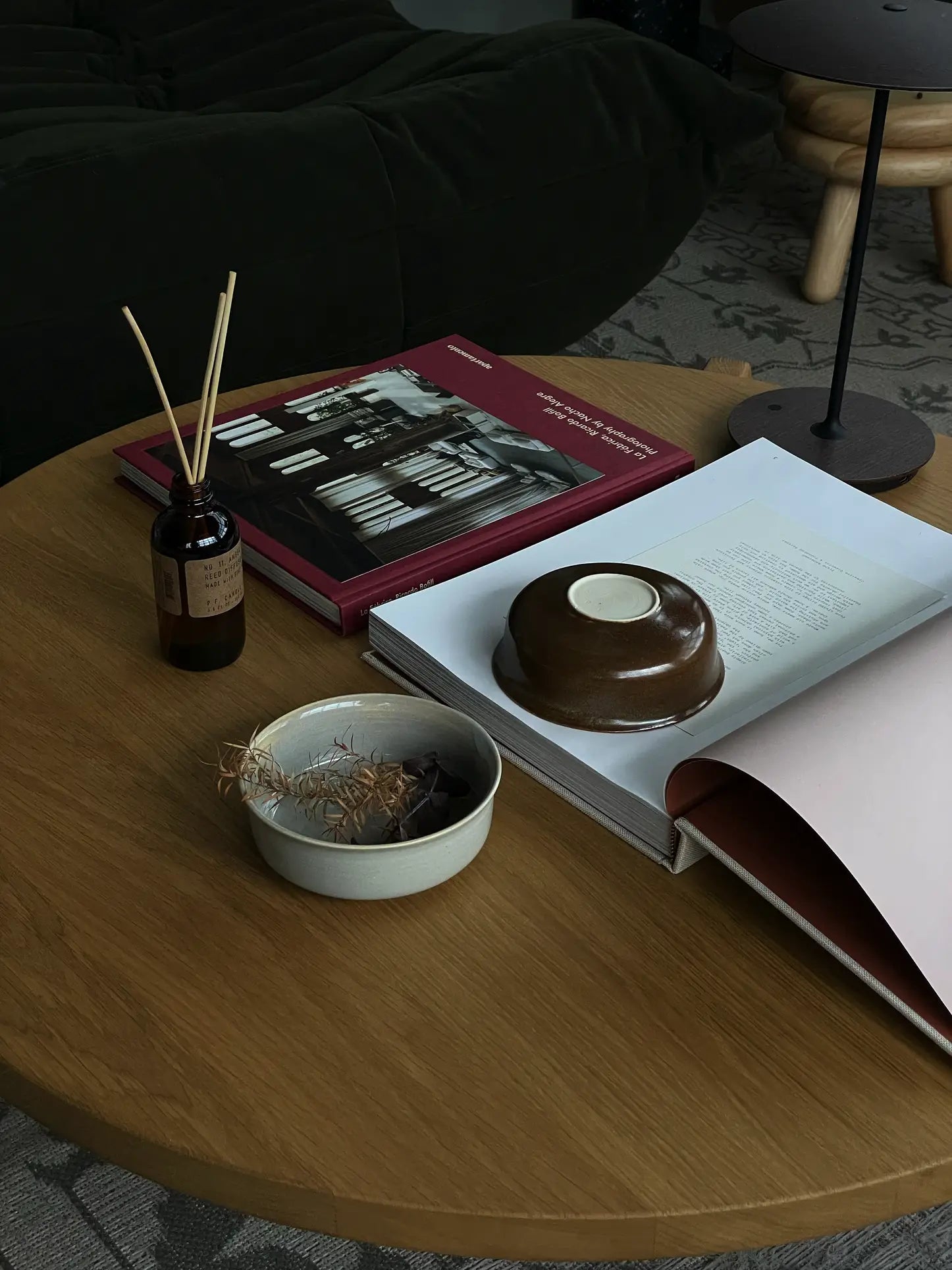 A wooden coffee table with a ves Aki Bowl, Set of 2 by ves in East Asian style filled with dried plants, an open and two closed books, a reed diffuser, a black lamp edge, and a dark sofa in the background.