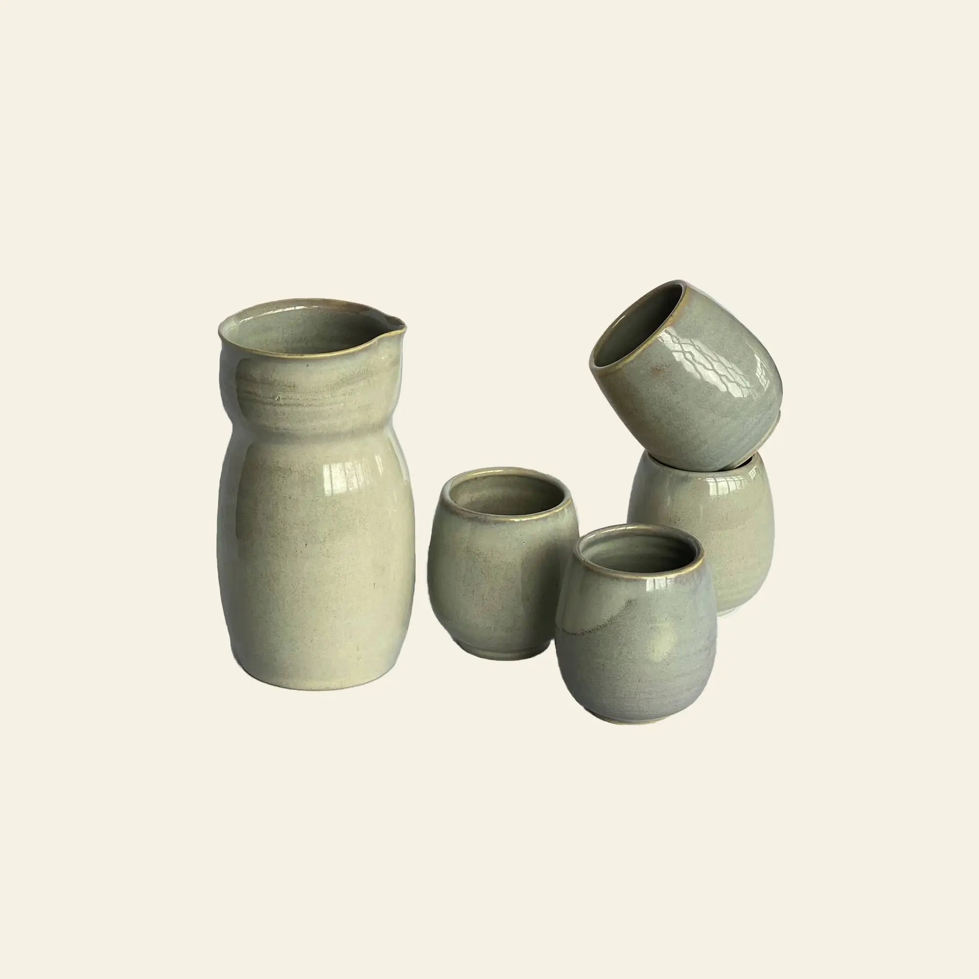The ves Aki Sake Set M by ves features four small, rounded ceramic cups and a matching jug in smooth light gray glaze, elegantly arranged on a light background with one cup stacked atop two others.