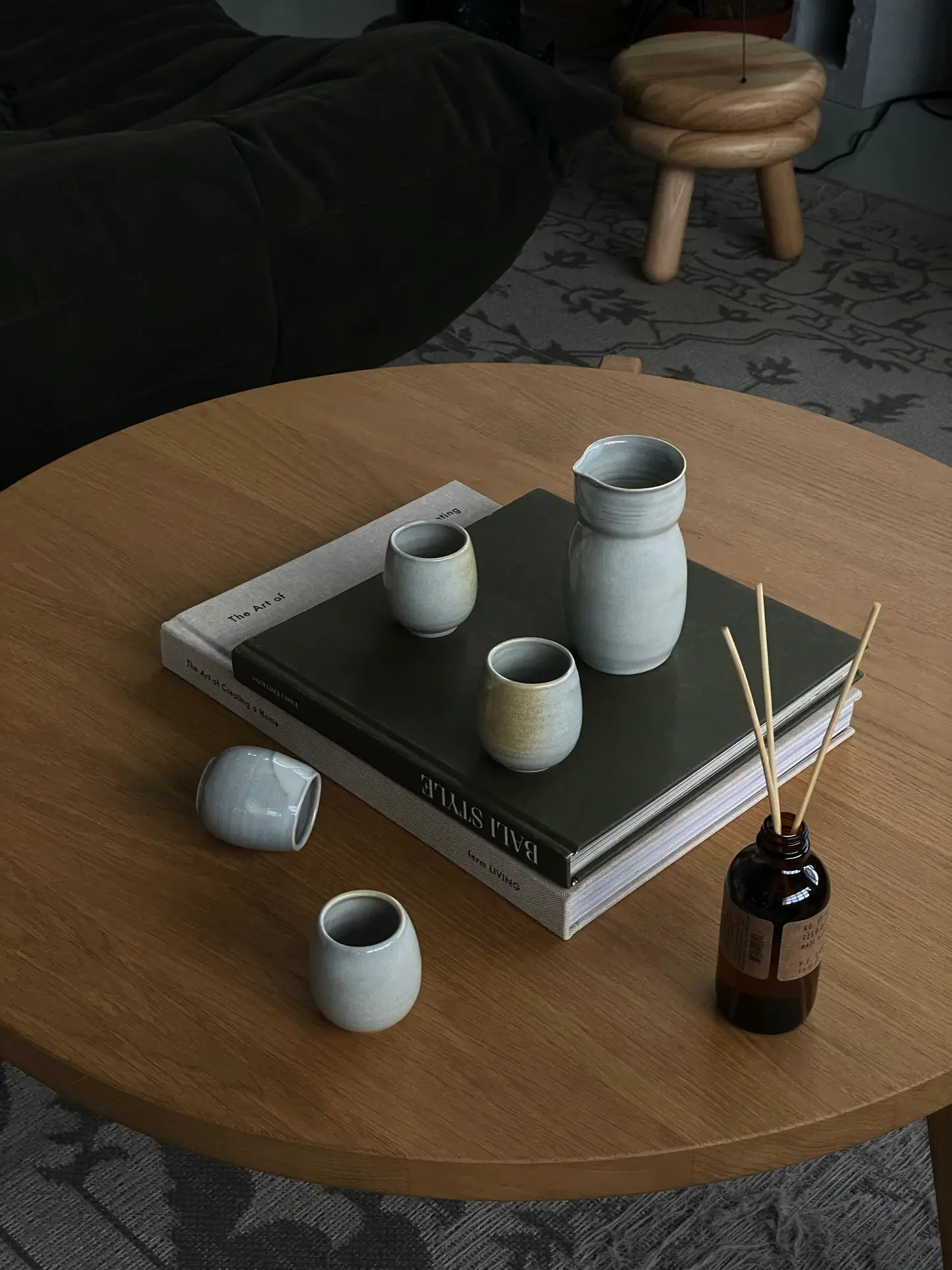 A round wooden table with handcrafted ceramic cups, a ves Aki Sake Set M pitcher, two stacked books, and a brown glass reed diffuser. In the background are a patterned rug, black sofa, and wooden stool.