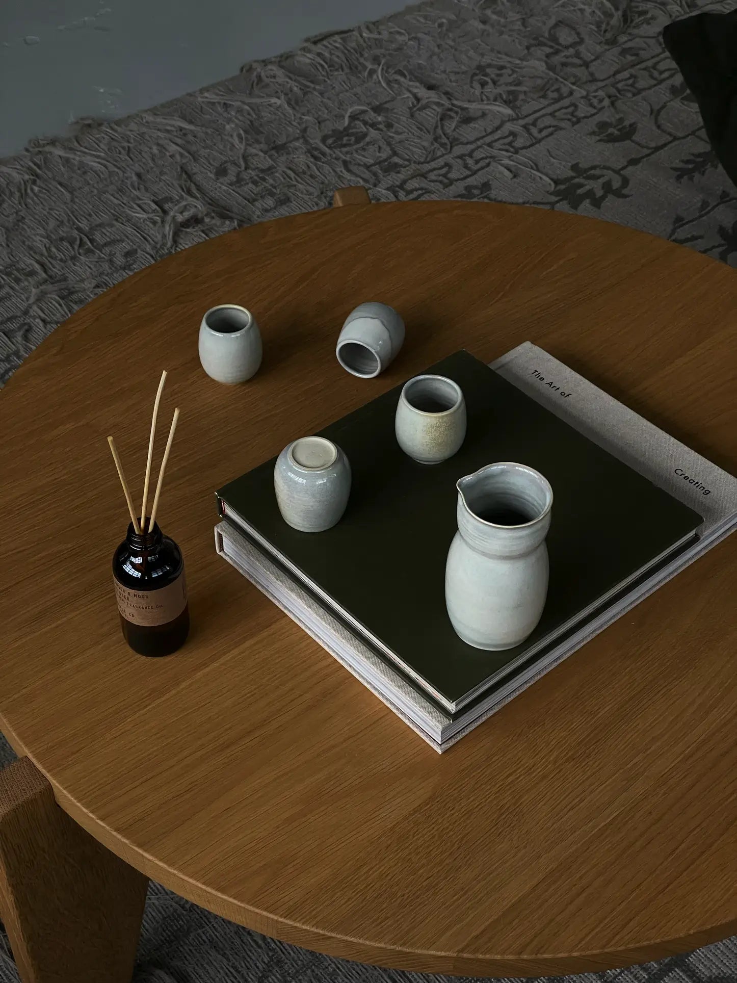 A round wooden coffee table holds several small ceramic vases, a ves Aki Sake Set M by ves, a reed diffuser, and two stacked books—one with a dark green cover. A patterned rug is partially visible underneath.
