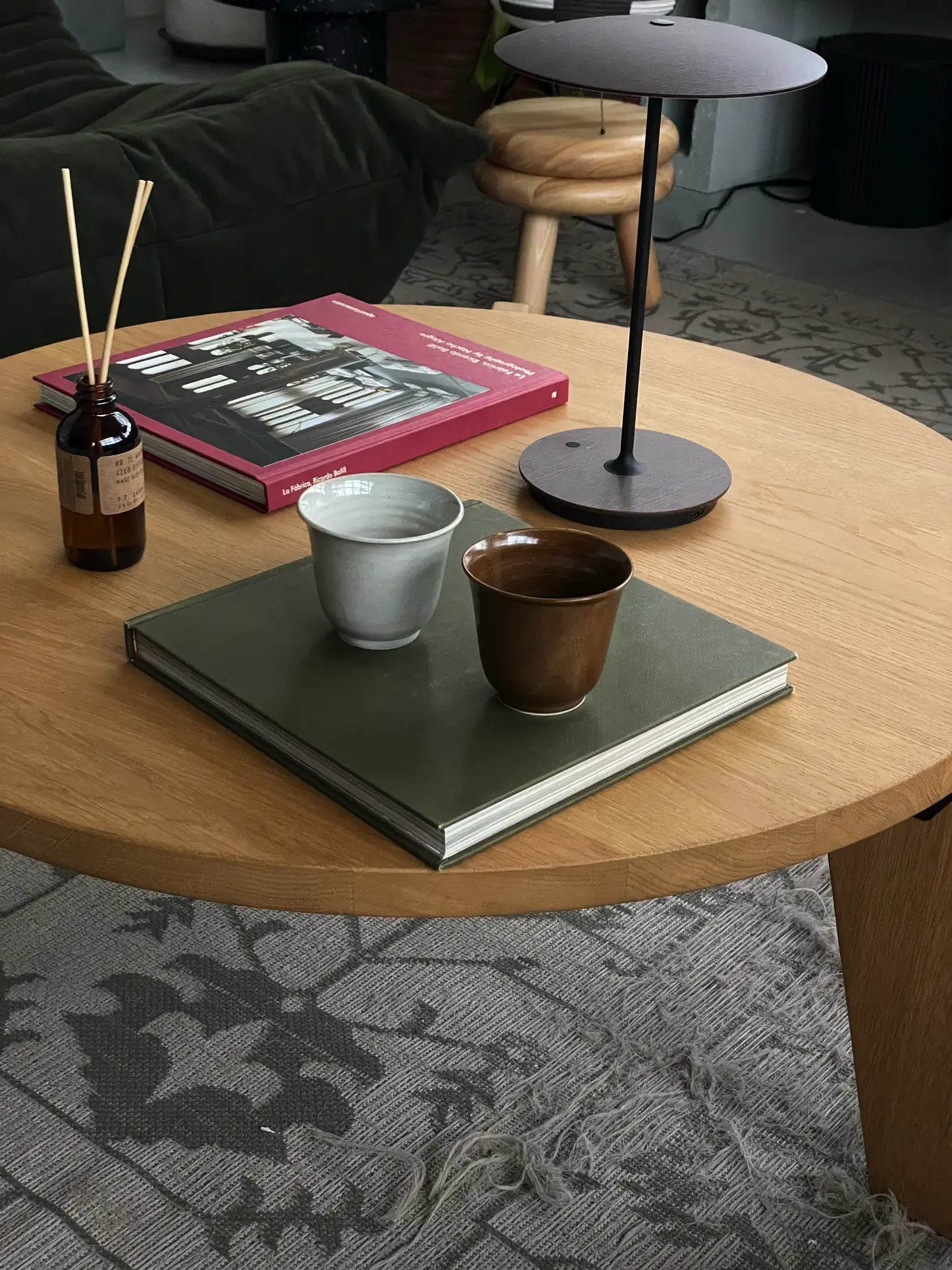 A handcrafted wooden coffee table displays a closed green book, ves Aki Teacup M, Set of 2 by ves, a reed diffuser, lamp, and red hardcover book. In the background: a sofa with blanket and woven stool from the elegant Aki Collection.