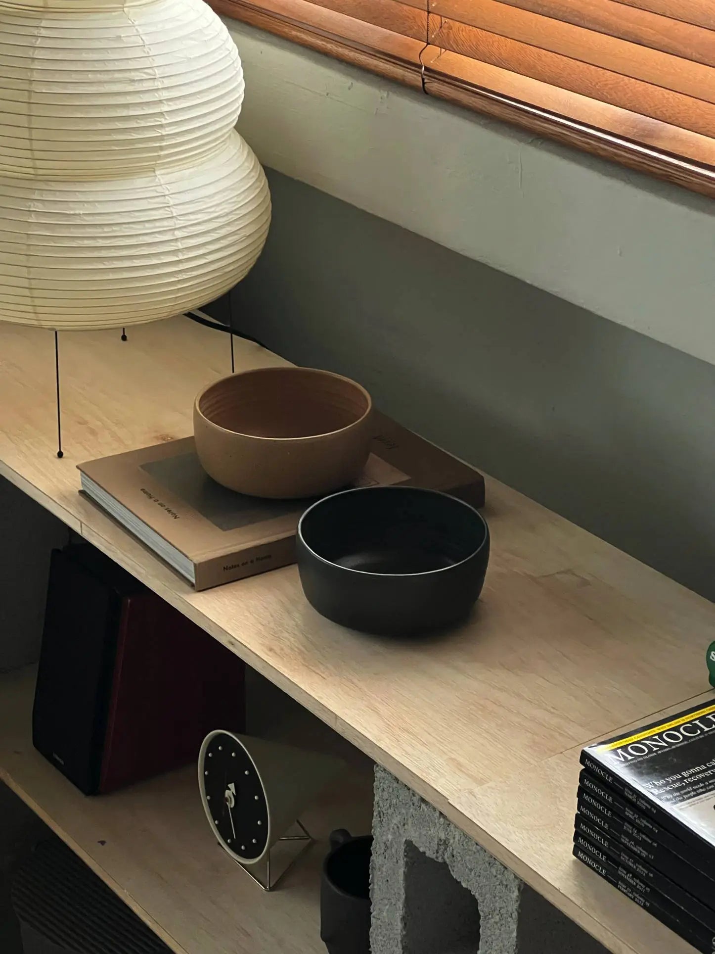 A light wooden desk features the ves Boro Ramen Bowl, Set of 2 by ves, stacked books, a modern white paper lamp, and magazines. Sunlight streams through wooden blinds above, casting soft shadows on the scene.