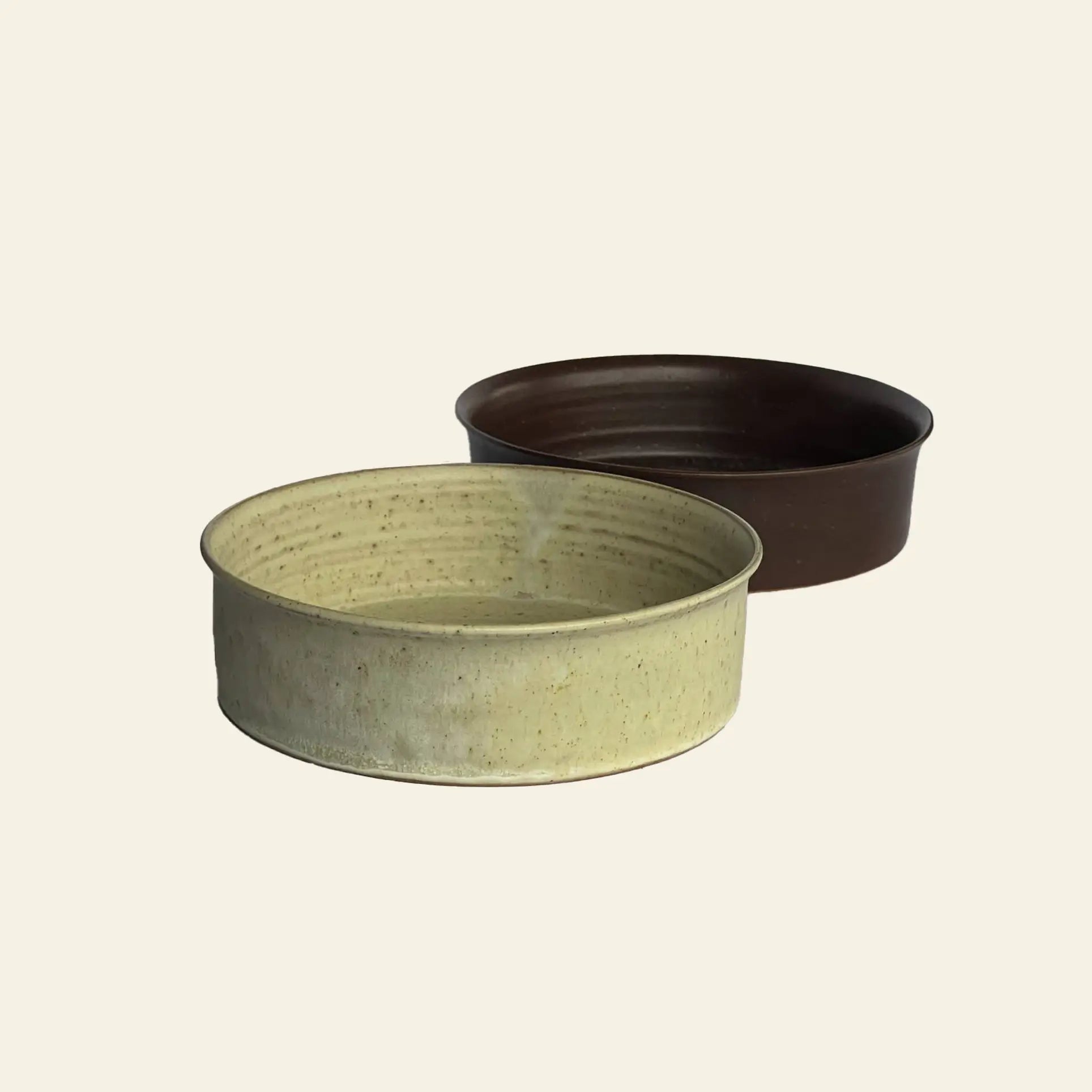 Two round ceramic bowls from the ves Kura Bowl M, Set of 2 are shown on a plain light background. The front bowl is light beige with a speckled finish, while the back one is solid dark brown, highlighting modern handcrafted design.