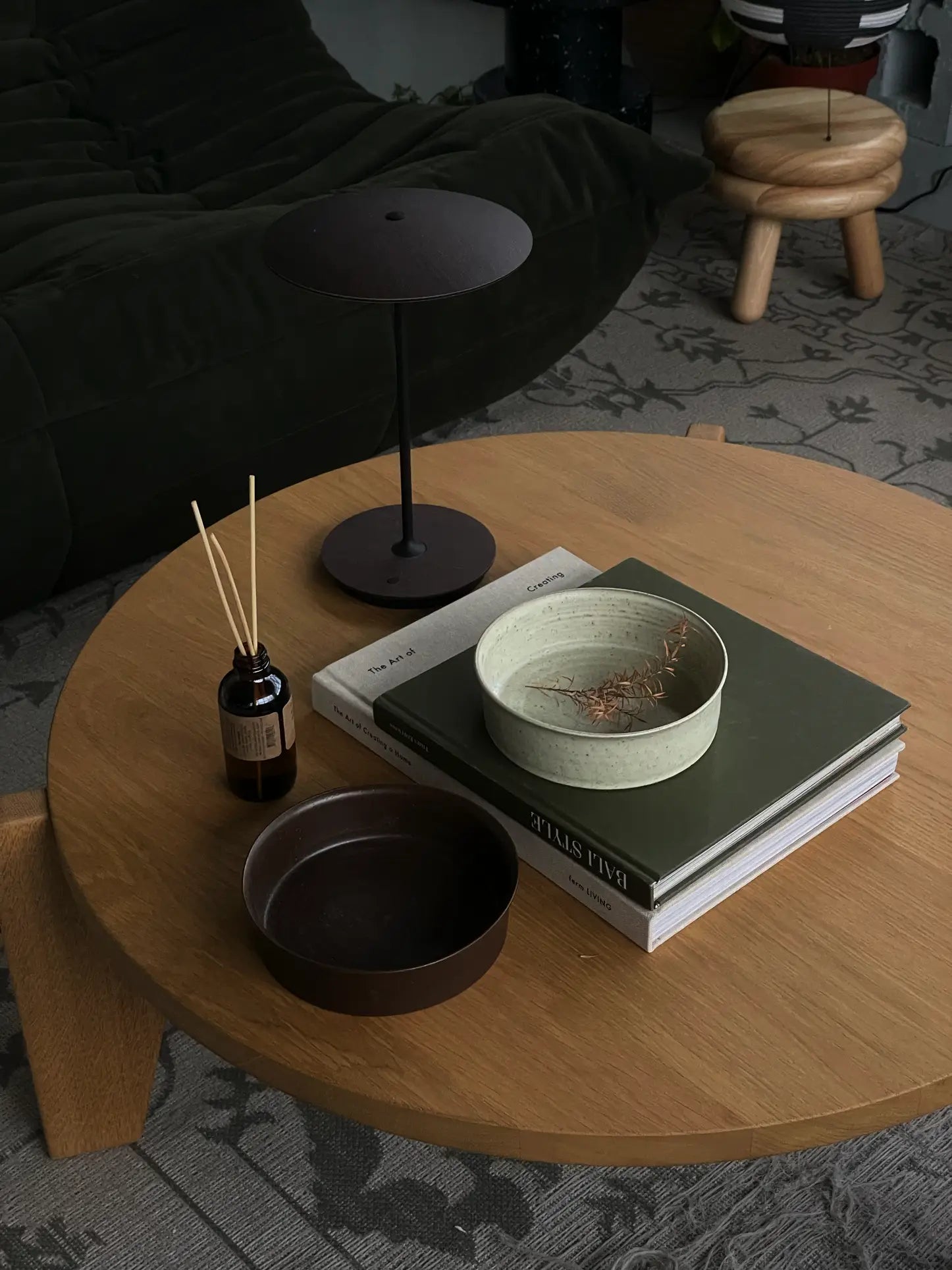 A round wooden coffee table holds a dark lamp, handcrafted ceramics like the ves Kura Bowl M, Set of 2 by ves, a reed diffuser, and stacked books in a cozy living room with a dark sofa and patterned rug in the background.