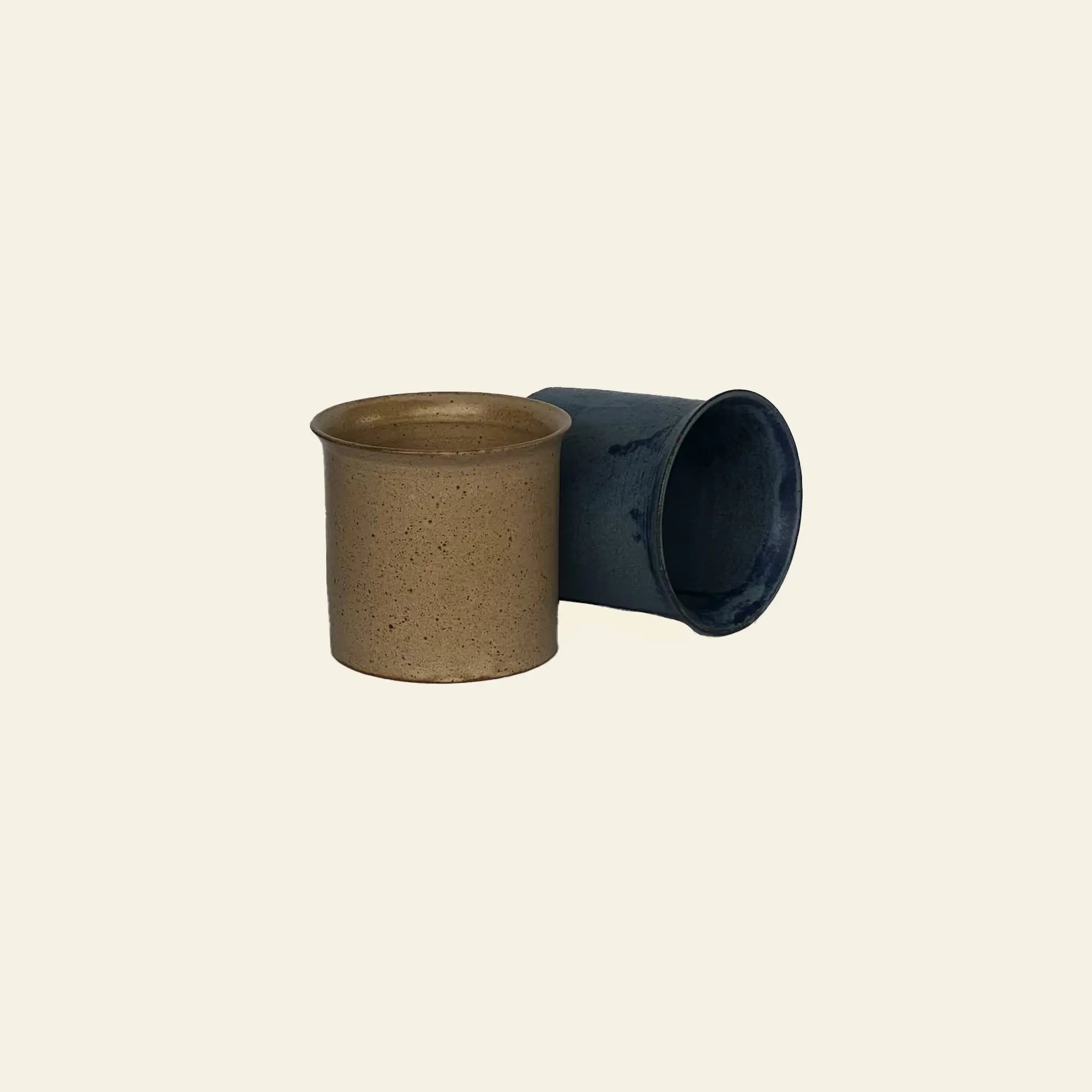 The ves Kura Cup Set of 2 by ves features one tan upright and one blue cylindrical cup, adding handcrafted ceramic charm to your decor, shown against a beige background.