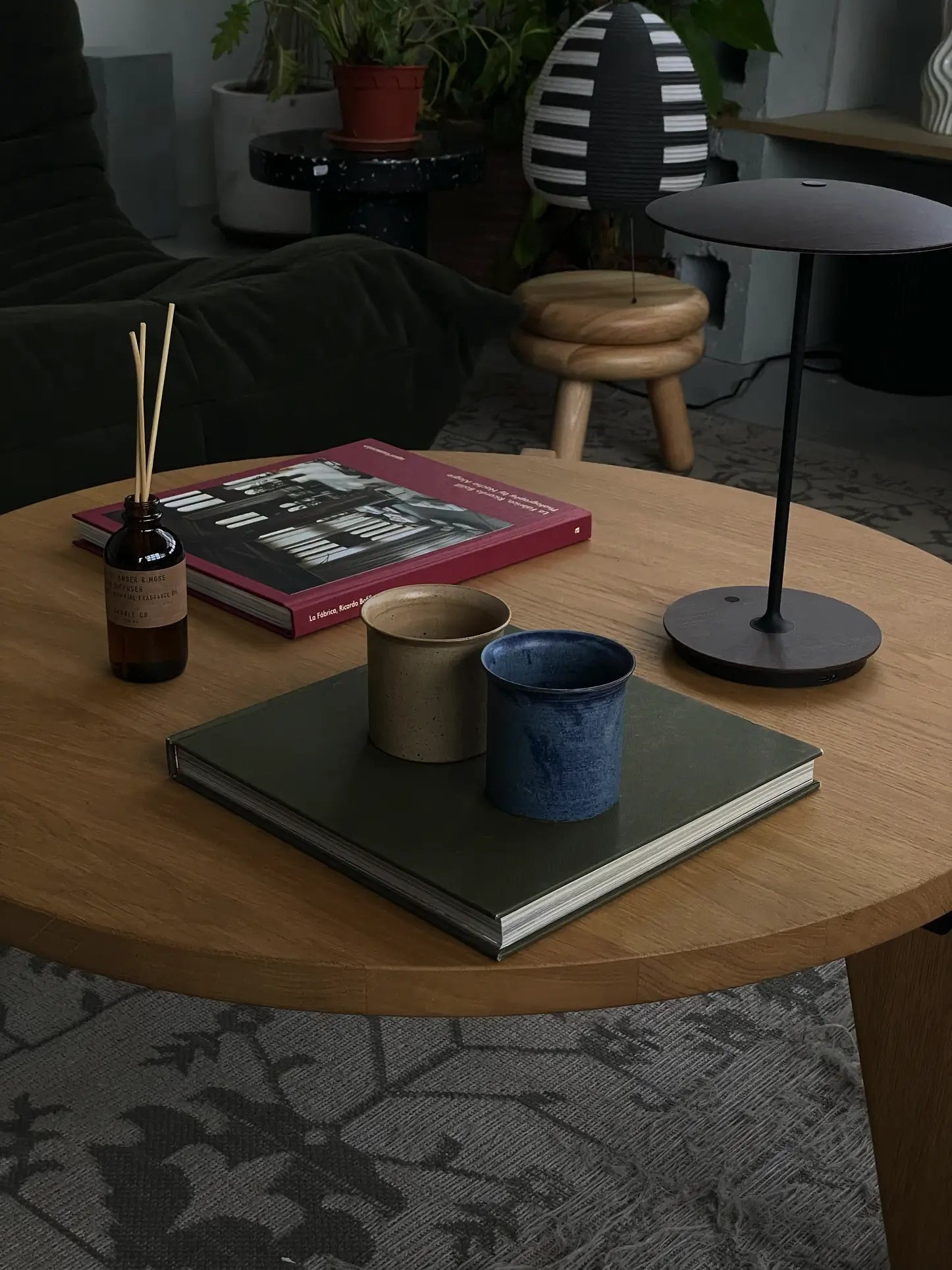 A round wooden coffee table from the ves Kura Collection displays a ves Kura Cup set of 2, a closed book, reed diffuser, and black lamp. In the background: a black chair, small stool, and potted plants complete the decor.