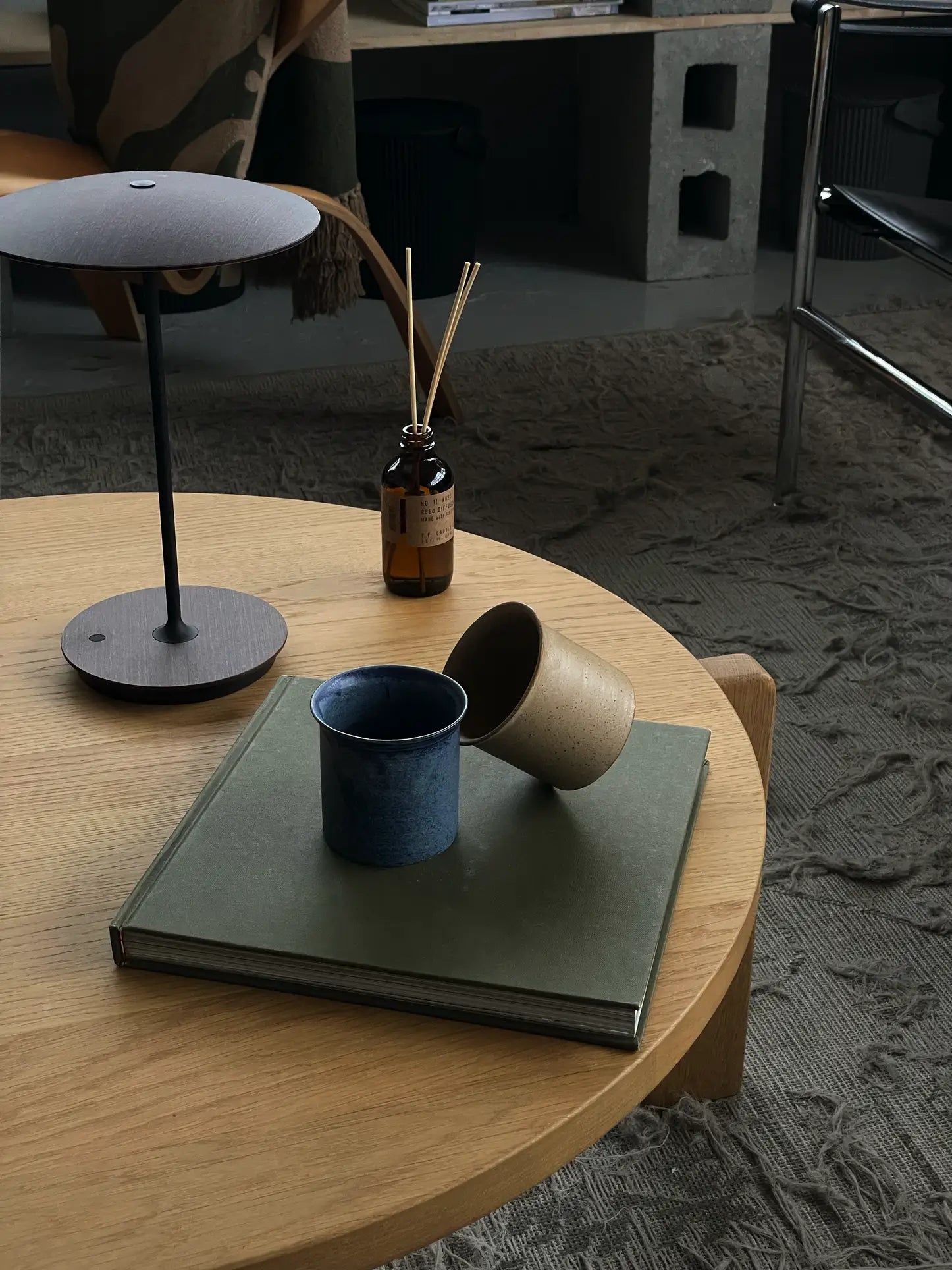 A round wooden coffee table displays a green book, the ves Kura Cup, Set of 2 by ves in blue and beige, a brown bottle with reed sticks, and a modern gold lamp. The background shows a textured rug and chic decor shelves.