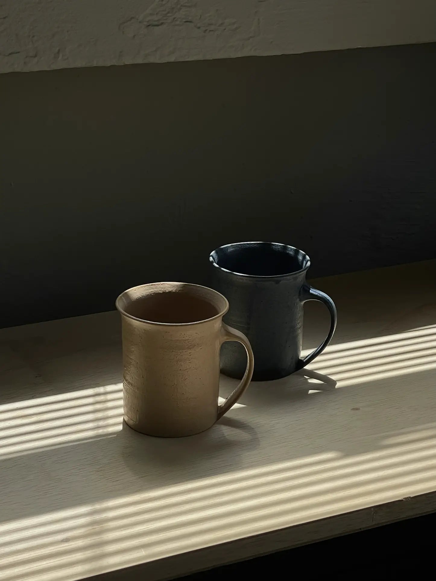 The ves Kura Mug, Set of 2 by ves features one beige and one dark blue ceramic mug with ergonomic handles, displayed side by side on a light wooden surface with sunlight casting striped shadows through blinds.