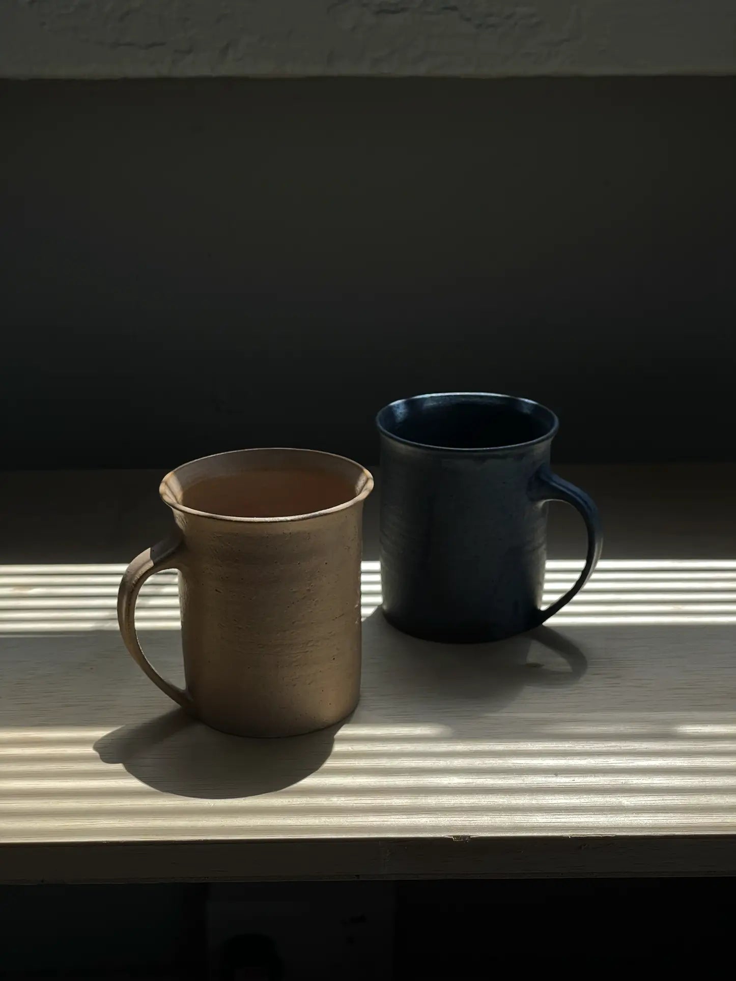 The ves Kura Mug Set of 2 by ves features handcrafted beige and dark blue ceramics with ergonomic handles, displayed side by side on a wooden surface beneath striped sunlight and soft shadows.