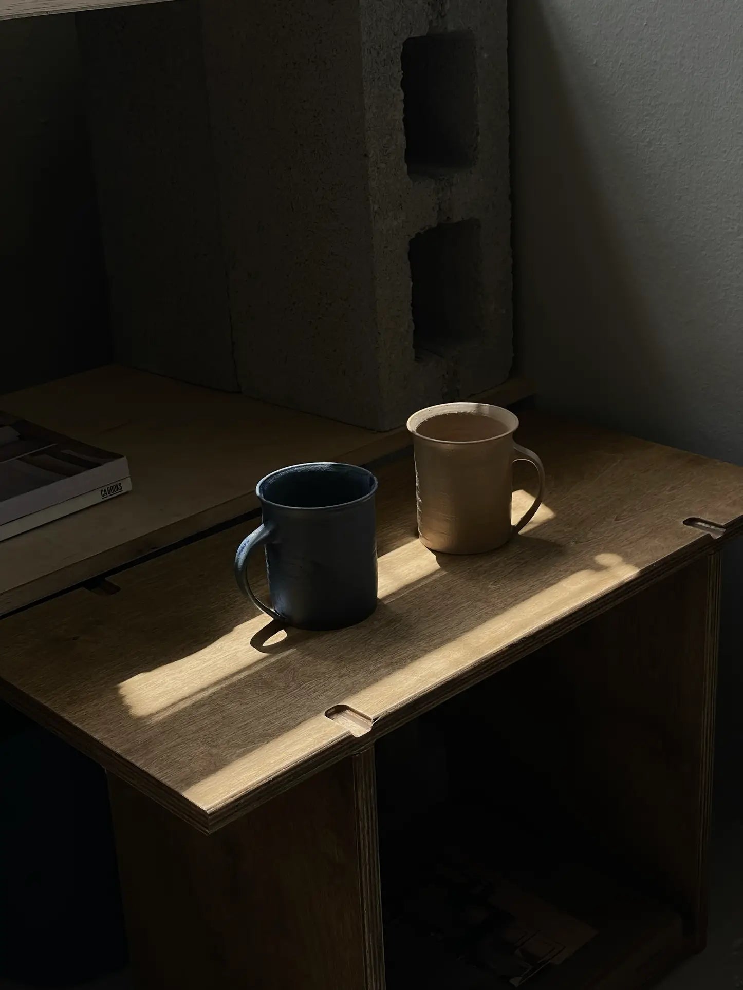 Two ves Kura Mugs from ves, one dark and one light, sit side by side on a wooden table with sunlight casting soft shadows. Concrete blocks and a closed book are in the background. Each mug in this set of 2 has an ergonomic handle.