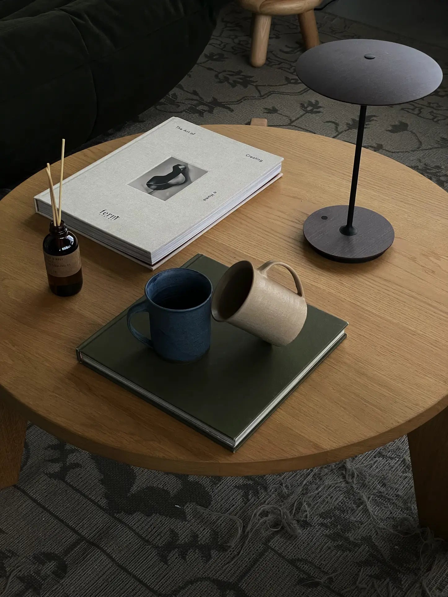 A round wooden coffee table holds the ves Kura Mug, Set of 2 by ves, each with ergonomic handles, a green book, a dark lamp, a reed diffuser, and a minimalist art magazine—all atop a patterned Kura Collection rug.