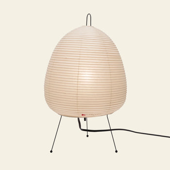 The Akari 1A table lamp by Akari features a Japanese-inspired ribbed, egg-shaped shade and three slim black legs, evoking classic Akari Light Sculptures, photographed against an off-white background.