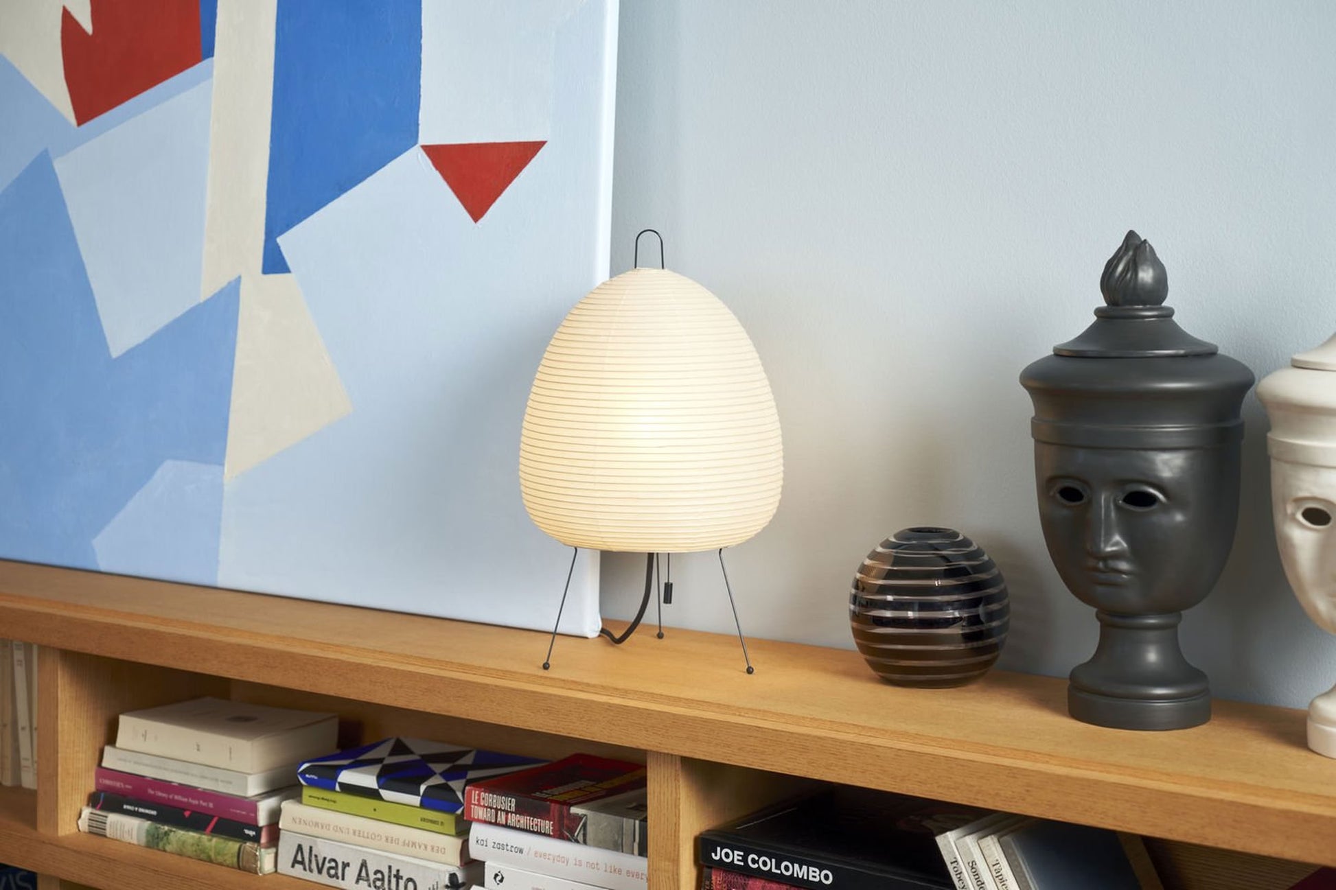 The Akari 1A table lamp by Akari, featuring a ribbed oval handmade paper shade inspired by Japanese lanterns, sits on a wooden shelf beside decor. Above it, an abstract geometric painting adds to the stylish display.