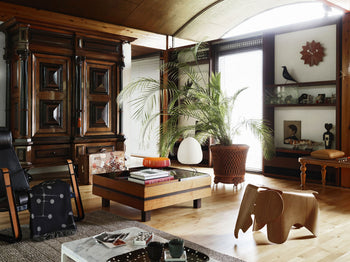 A stylish living room with wooden furniture, lush plants, books on a coffee table, a rocking chair, and the Akari 1A Japanese lantern by Akari made of handmade paper. Sunlight streams through large windows onto an art-filled display cabinet.