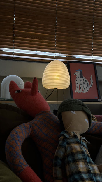 Two plush toys—a red animal with long ears and a plaid-shirt doll—sit on a couch. Behind them are an Akari 1A lantern by Akari, a framed picture of a spotted dog, and wooden blinds.