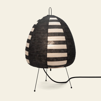 The Akari 1AS table lamp by Akari, inspired by Akari Light Sculptures, features a black and white striped paper shade and slim angled metal legs. It glows softly to highlight modern design and Japanese craftsmanship, with a side-extending power cord.
