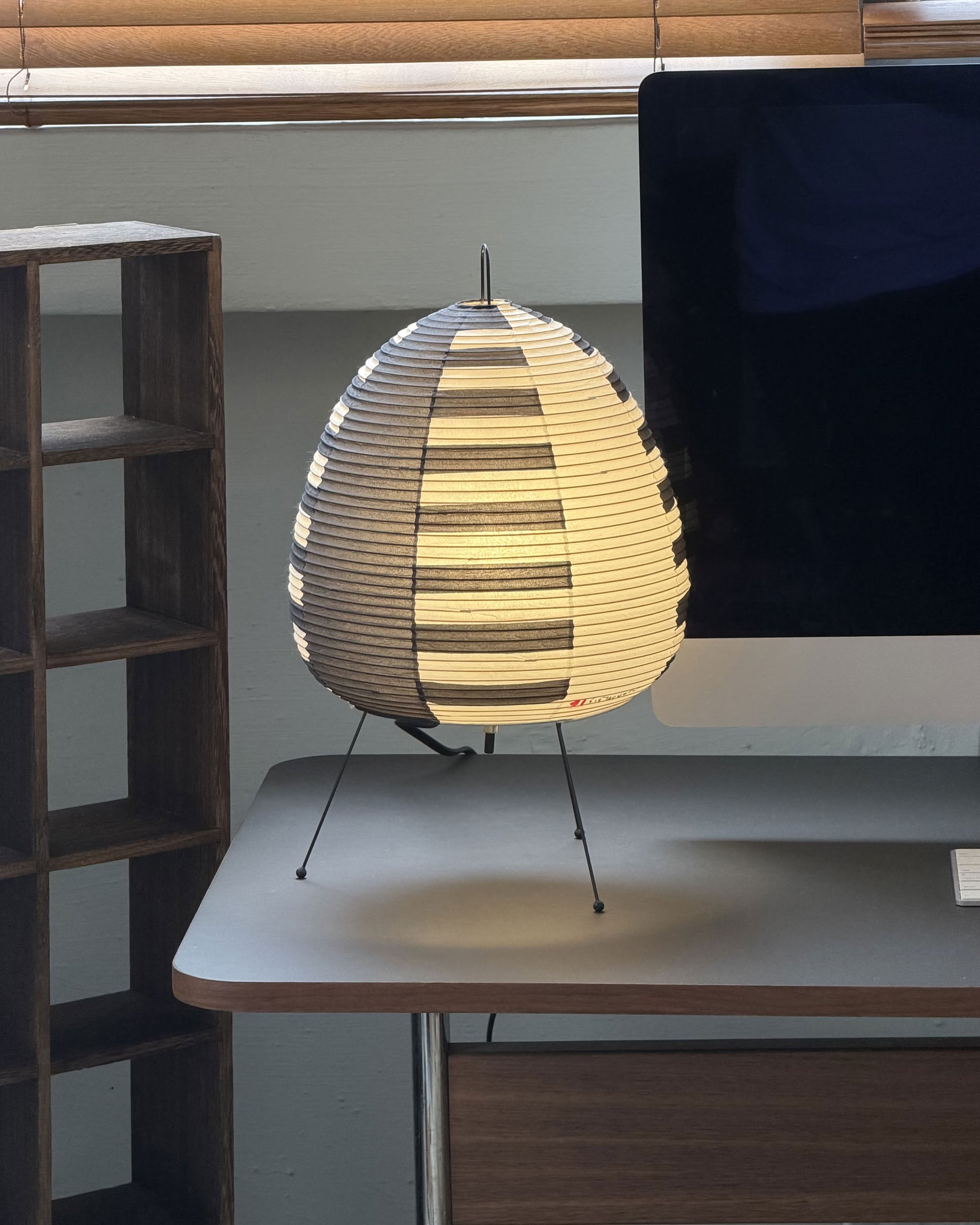 An Akari 1AS paper lantern by Akari, featuring a striped, oval design handcrafted from washi paper, stands on three metal legs atop a desk near a computer monitor and wooden shelf, with window blinds above.