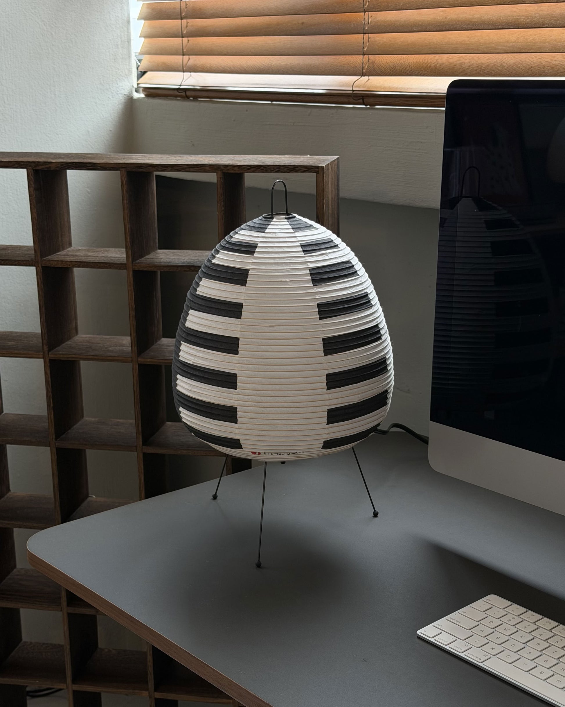 The Akari 1AS by Akari, a modern table lamp with a piano keyboard-style black and white shade inspired by Japanese lanterns, is placed on a gray desk beside a computer monitor, keyboard, and wooden shelving under window blinds.