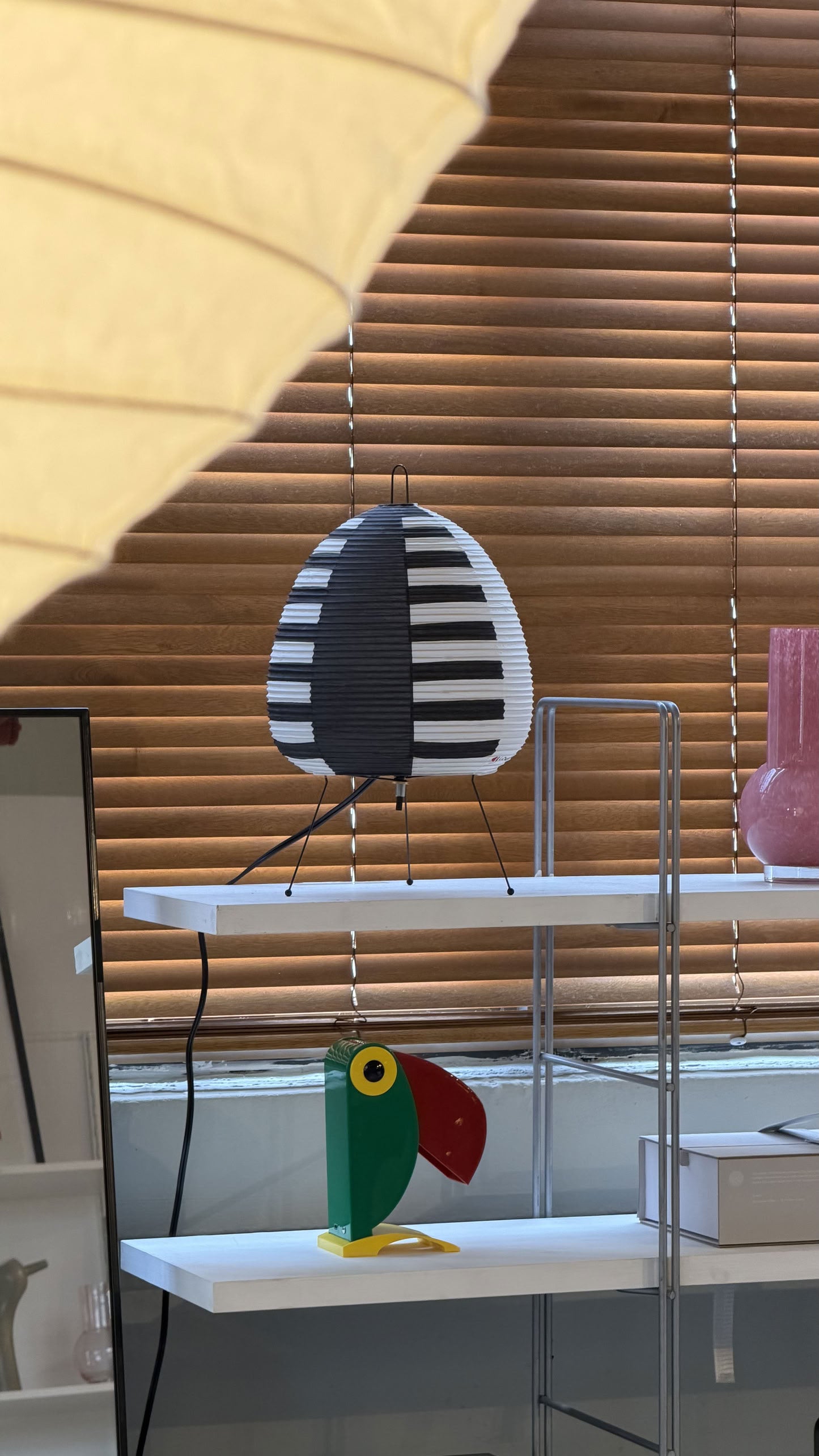 The Akari Akari 1AS table lamp, inspired by Japanese paper lanterns, sits on a white shelf in front of wooden blinds, accompanied by a colorful bird figurine and a pink vase.