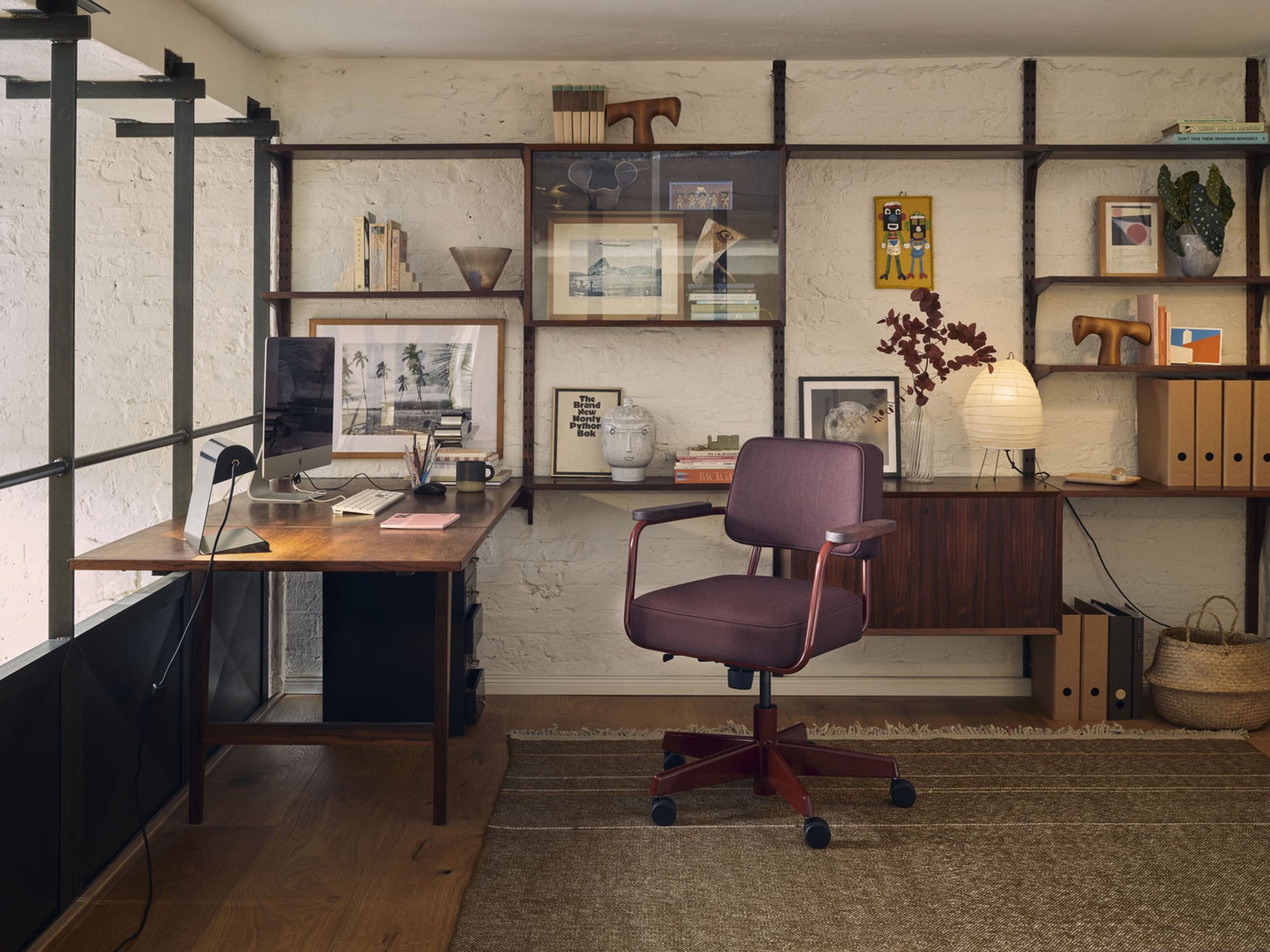 A modern home office with a burgundy chair, wooden desk, shelves for books and decor, and a neutral rug features the Akari 1N lamp by Akari—a Japanese paper lantern handcrafted in Japan—casting a soft glow on the white brick wall.