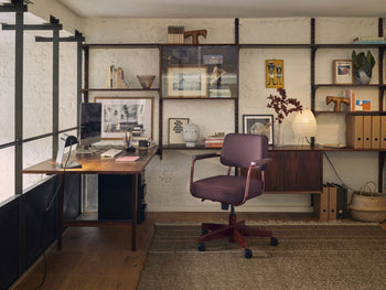 A modern home office with a burgundy chair, wooden desk, shelves for books and decor, and a neutral rug features the Akari 1N lamp by Akari—a Japanese paper lantern handcrafted in Japan—casting a soft glow on the white brick wall.