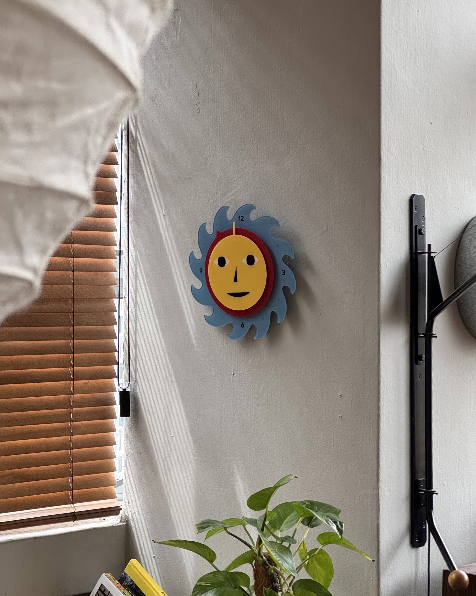 The Areaware Everybody Clock by Areaware, featuring a cheerful yellow face, adds a vibrant touch to the light gray wall near a window with wooden blinds and a green plant on the table below.